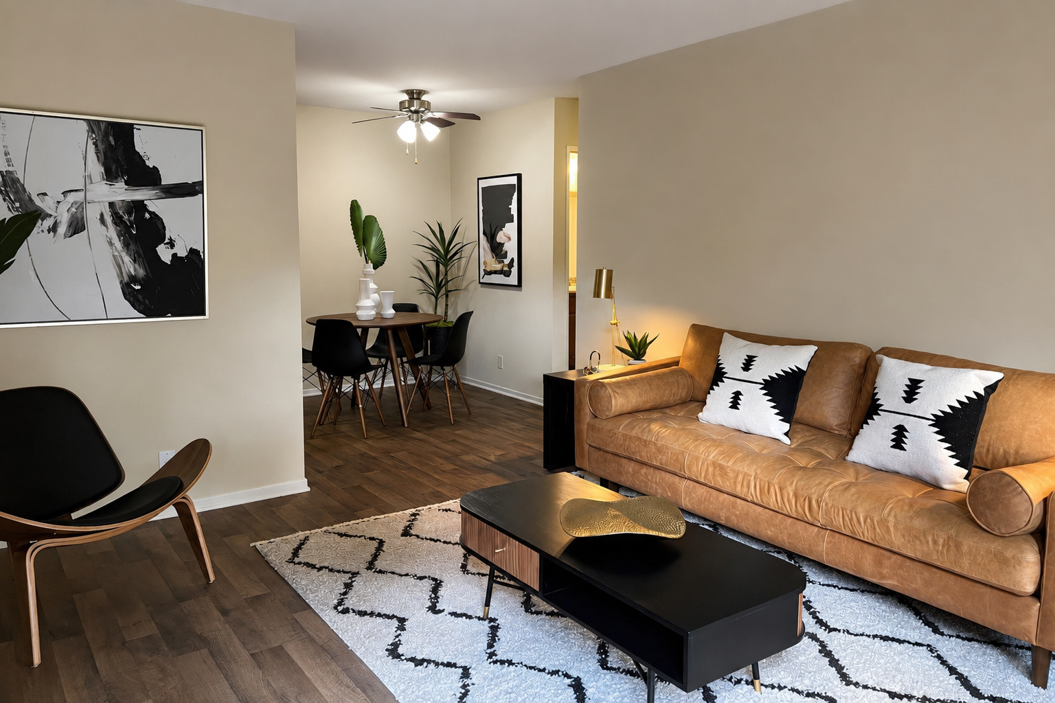 A stylish living room featuring a tan leather sofa with decorative cushions, a black coffee table on a patterned rug, and a wooden chair. In the background, there is a dining area with a table and chairs, along with indoor plants and wall art, creating a modern, inviting atmosphere.