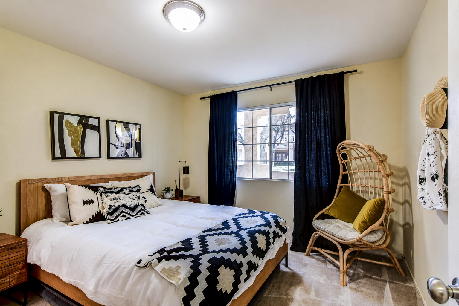 A cozy bedroom featuring a queen-sized bed with patterned black and white bedding, wooden nightstands on either side, and decorative wall art. A large window with dark curtains allows natural light, while a rattan chair with a cushion adds a comfortable seating option in the corner.