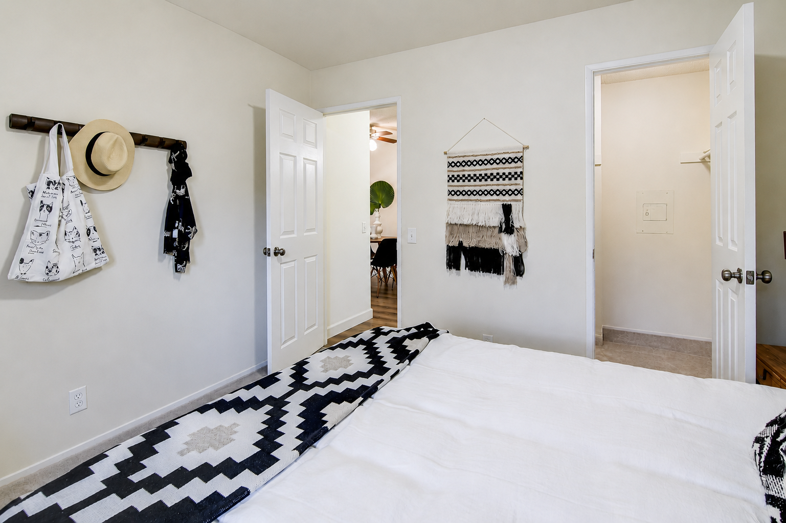 A well-decorated bedroom featuring a large bed with a white duvet. The walls are light-colored, and there are two open doors leading to other rooms. A black and white patterned rug is on the floor, with a hanging wall decor piece. A hat and bags are neatly arranged on a hook by the door.