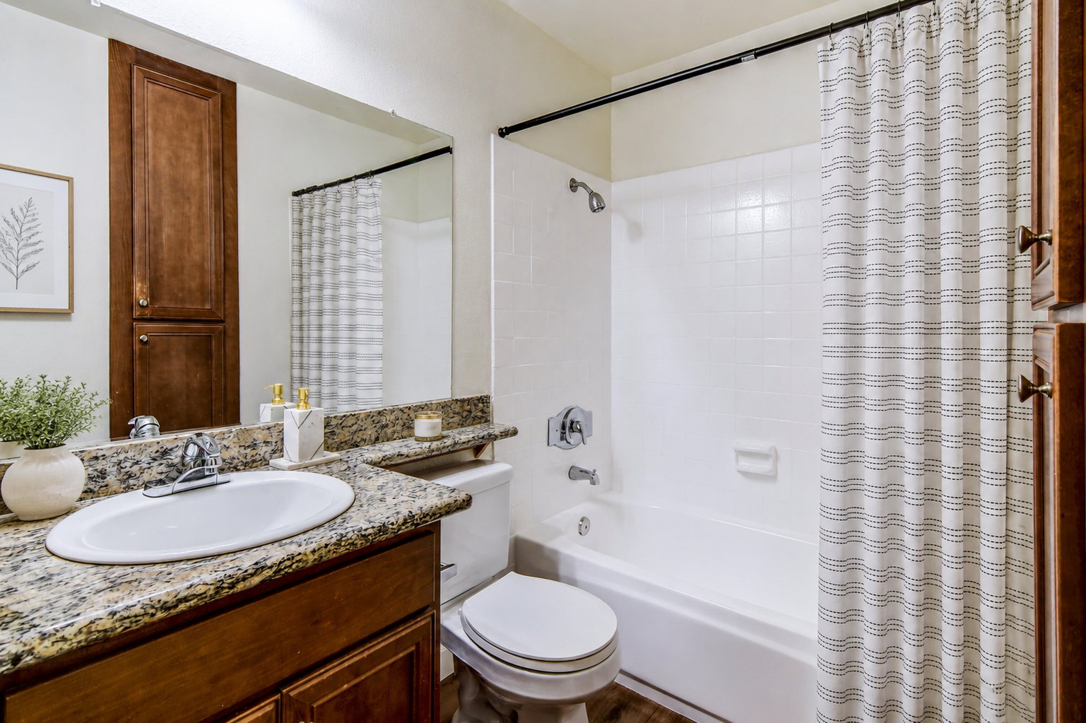 A clean and modern bathroom featuring a white bathtub and shower combination, a vanity with a round sink and granite countertop, a mirror above the sink, and a decorative plant. The walls are light-colored, and a patterned shower curtain is hanging beside the bathtub. Wooden cabinets and a towel rack complete the design.
