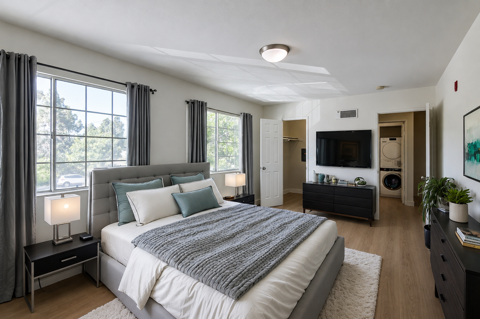 A modern bedroom featuring a king-sized bed with a gray and white color scheme, decorative pillows, and a cozy blanket. Two bedside tables with lamps are on either side of the bed. There is a large window letting in natural light, a mounted TV on the wall, and a closet in the background.