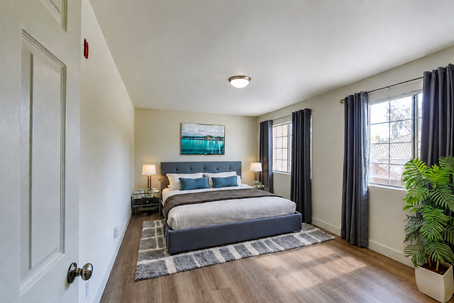 A spacious, well-lit bedroom featuring a large bed with blue pillows, bedside tables with lamps, a piece of artwork depicting a serene landscape, and large windows with curtains. A potted plant adds a touch of greenery to the room, which has wooden flooring and neutral-colored walls.