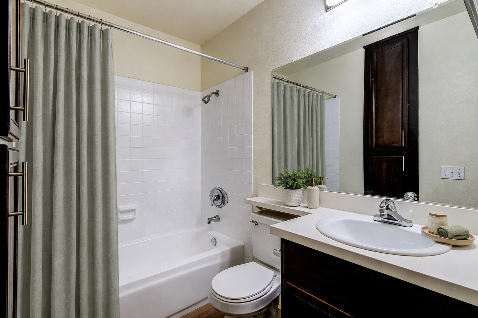 A modern bathroom featuring a white bathtub with a shower, light green curtains, a large mirror above the sink, and dark wooden cabinets. The countertop has a small plant and a folded towel. The overall color scheme is neutral and bright, creating a clean and inviting atmosphere.