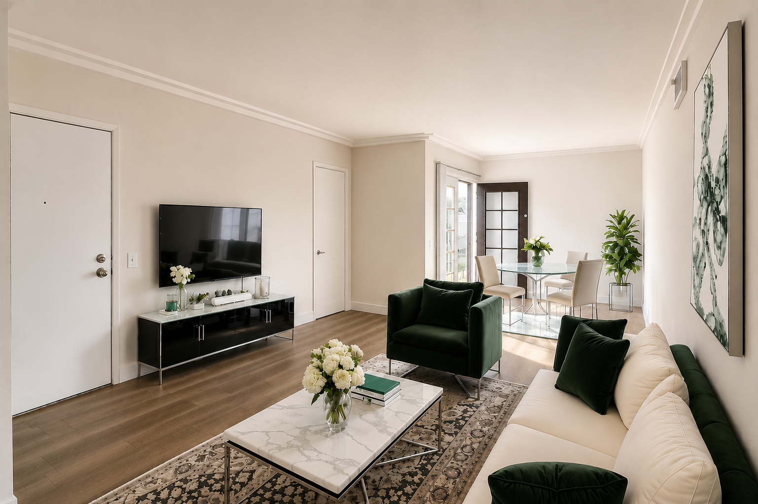Modern living room featuring a neutral color palette with light beige walls and hardwood floors. A black TV stand holds a flat-screen TV, and a white marble coffee table sits centrally. Stylish green sofas and a dining area with a round table and chairs are visible, along with indoor plants and decorative floral arrangements.
