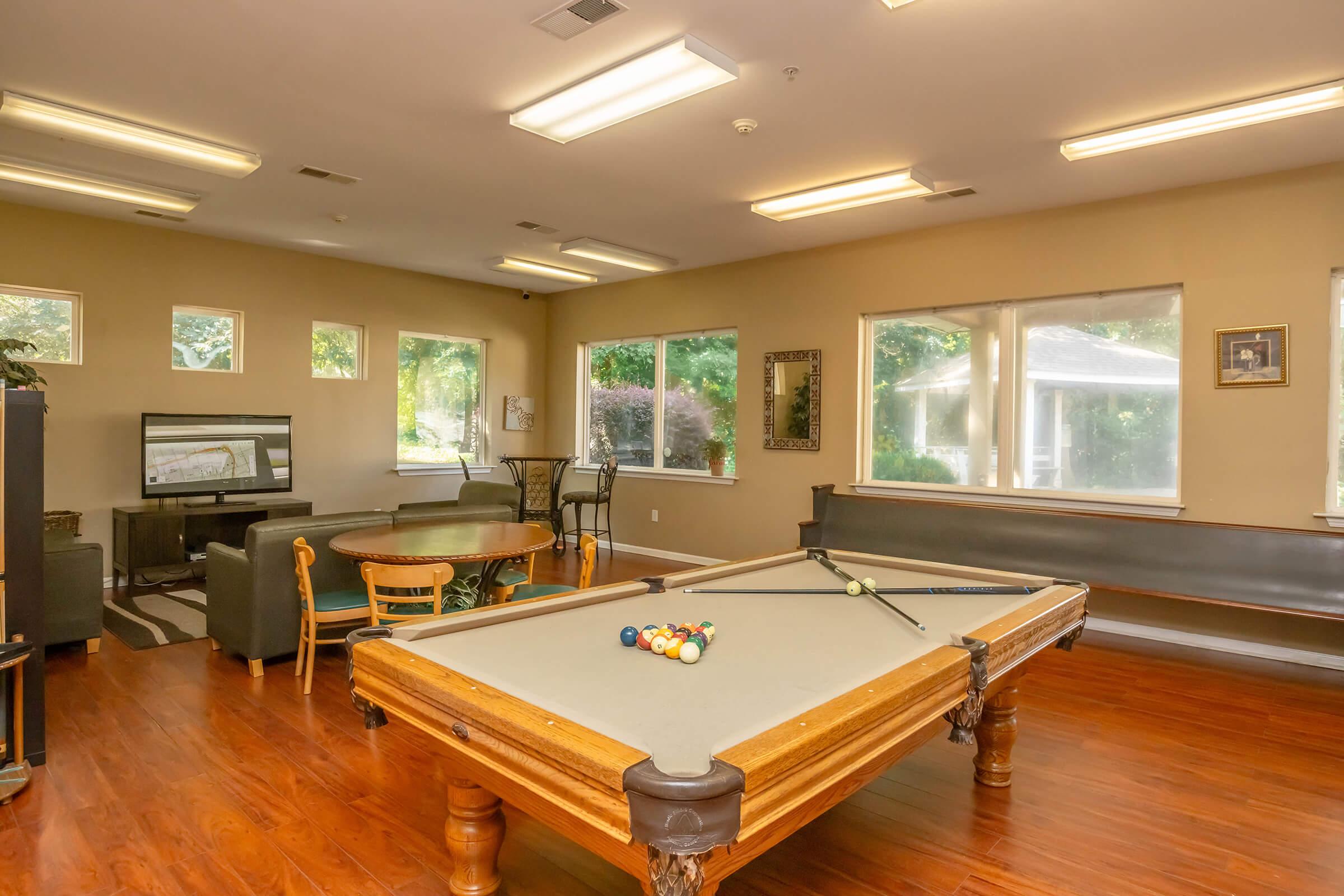 A well-lit recreation room featuring a pool table in the foreground, surrounded by a seating area with sofas and a dining table. Large windows allow natural light to fill the space, creating a welcoming atmosphere. The walls are adorned with framed pictures, enhancing the inviting decor.