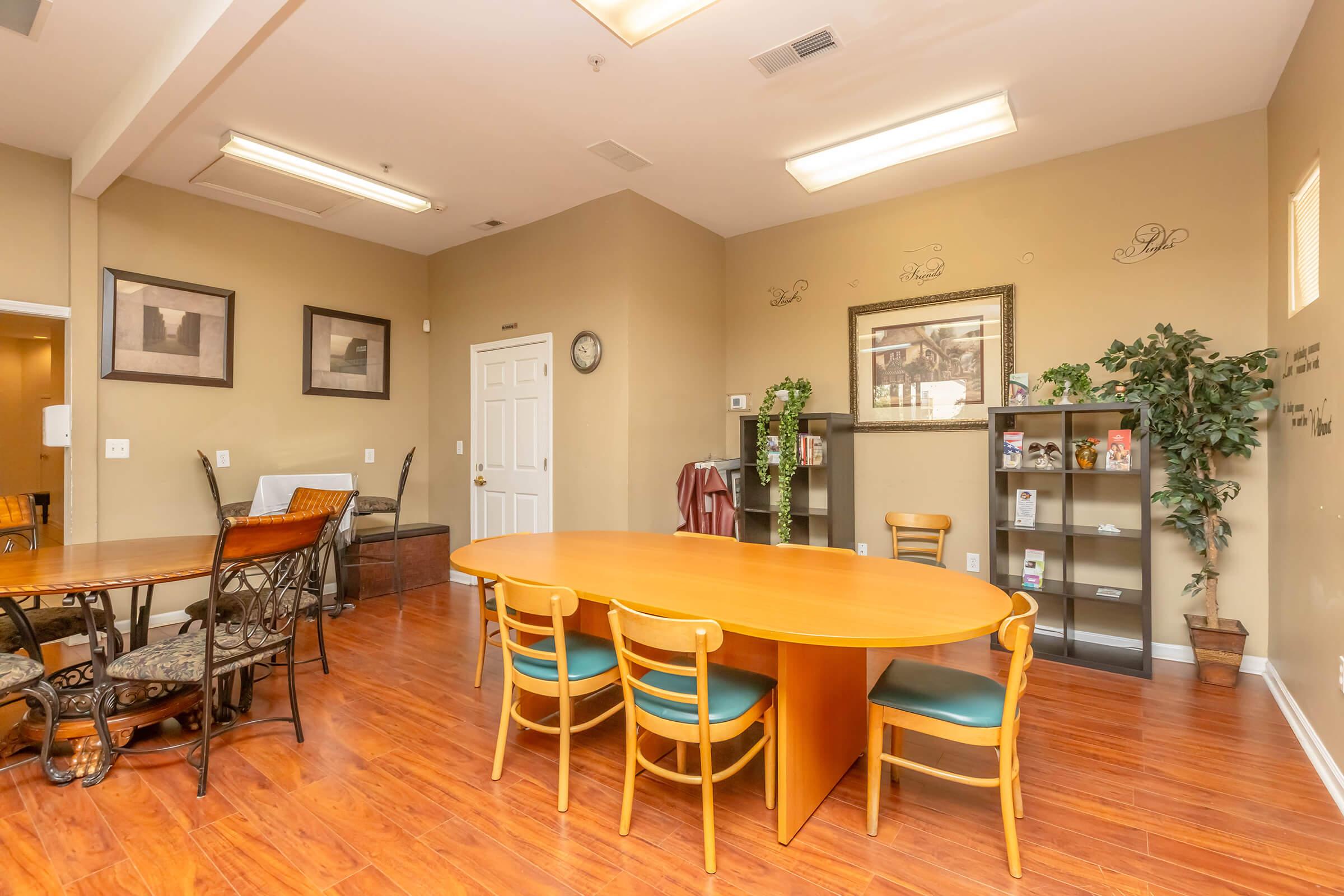 Interior of a room featuring a round wooden table with several chairs, a small dining table with metal chairs, and a decorative plant. There are framed artworks on the wall, a clock, and informational brochures on a stand. The floor is wooden and the walls have a warm beige color, creating a welcoming atmosphere.