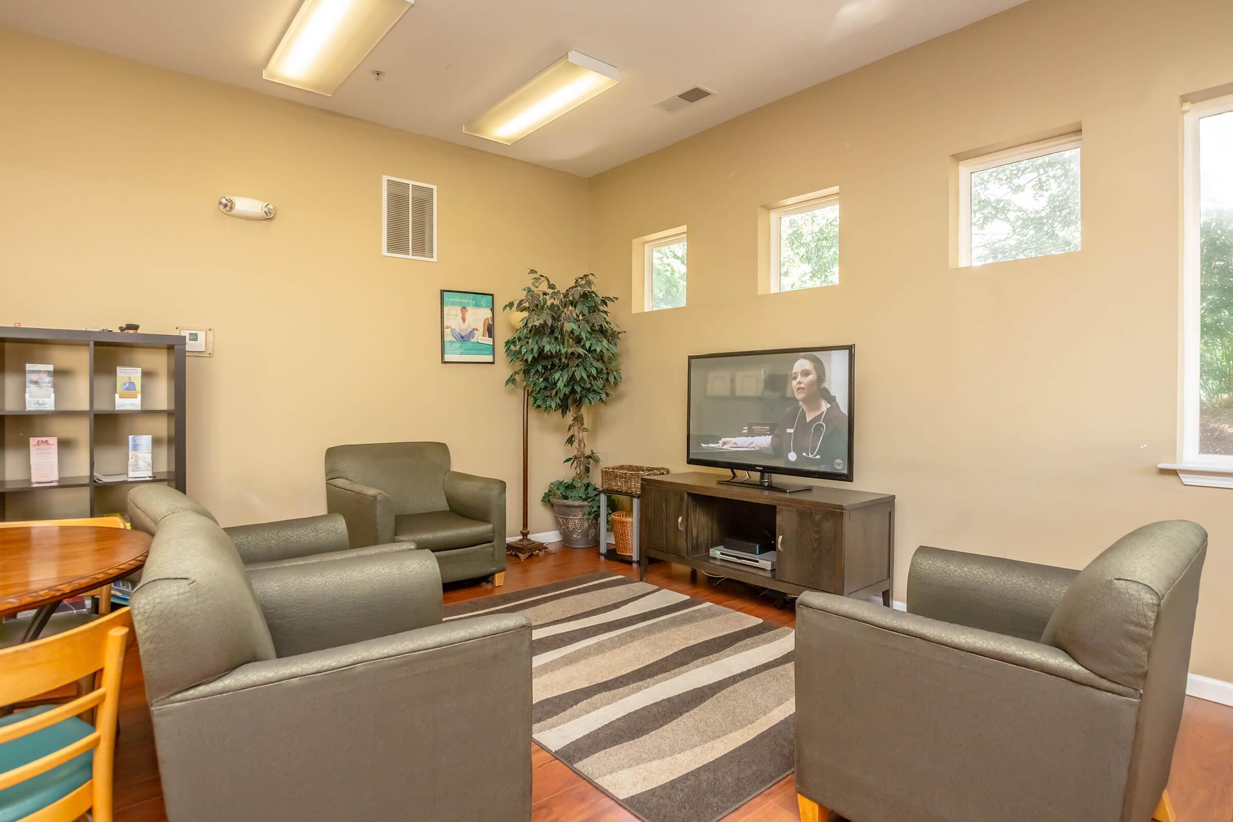 A cozy living room with four armchairs arranged around a coffee table. A television stands on a wooden cabinet, displaying a program. There are windows letting in natural light, and a potted plant adds a touch of greenery to the beige walls and wooden floor.
