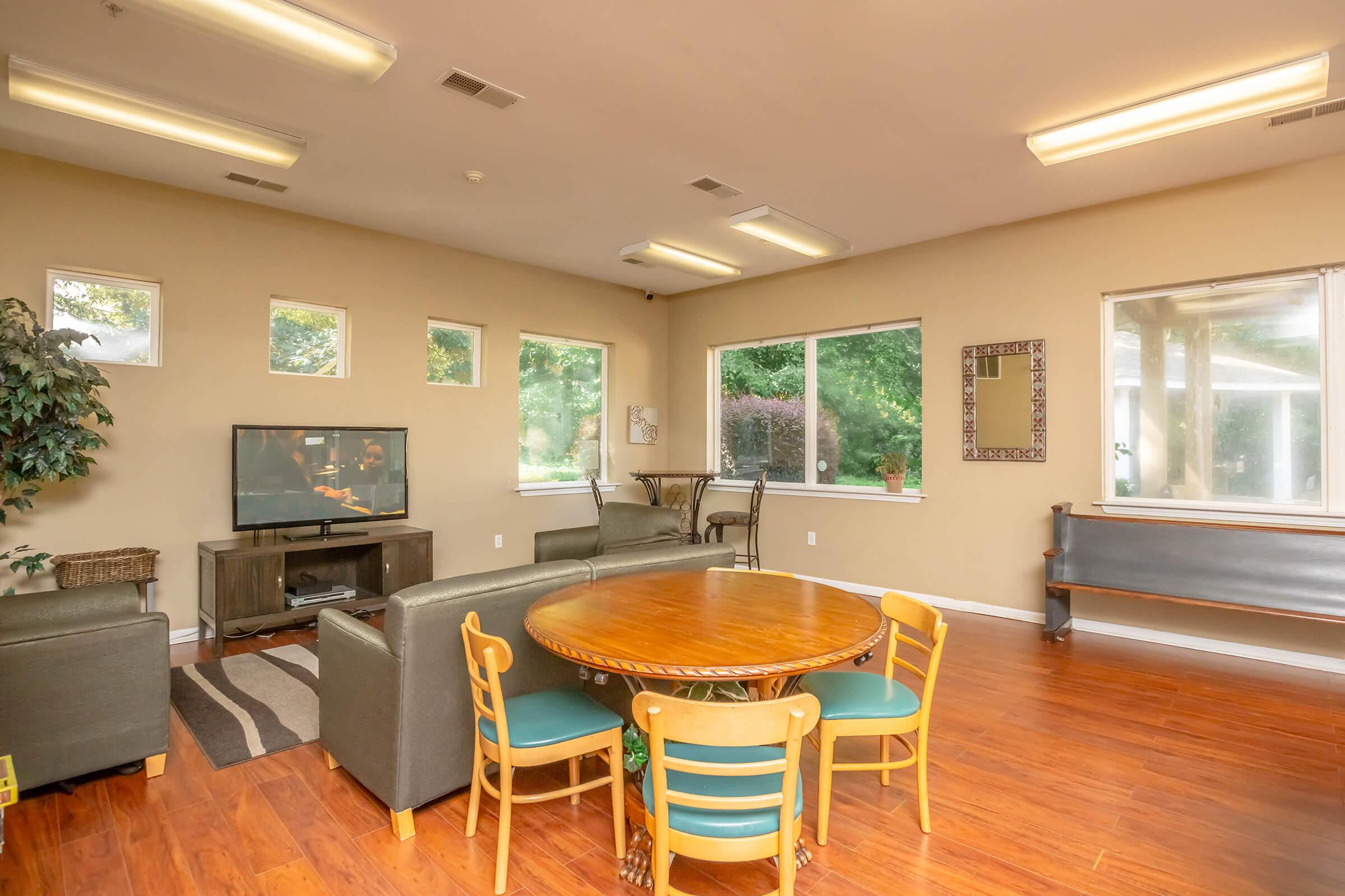 A cozy living space featuring a circular wooden dining table surrounded by chairs, green sofas, and a television. Light-colored walls and large windows allow natural light to fill the room, with potted plants adding a touch of greenery. The flooring is wooden, creating a warm and inviting atmosphere.