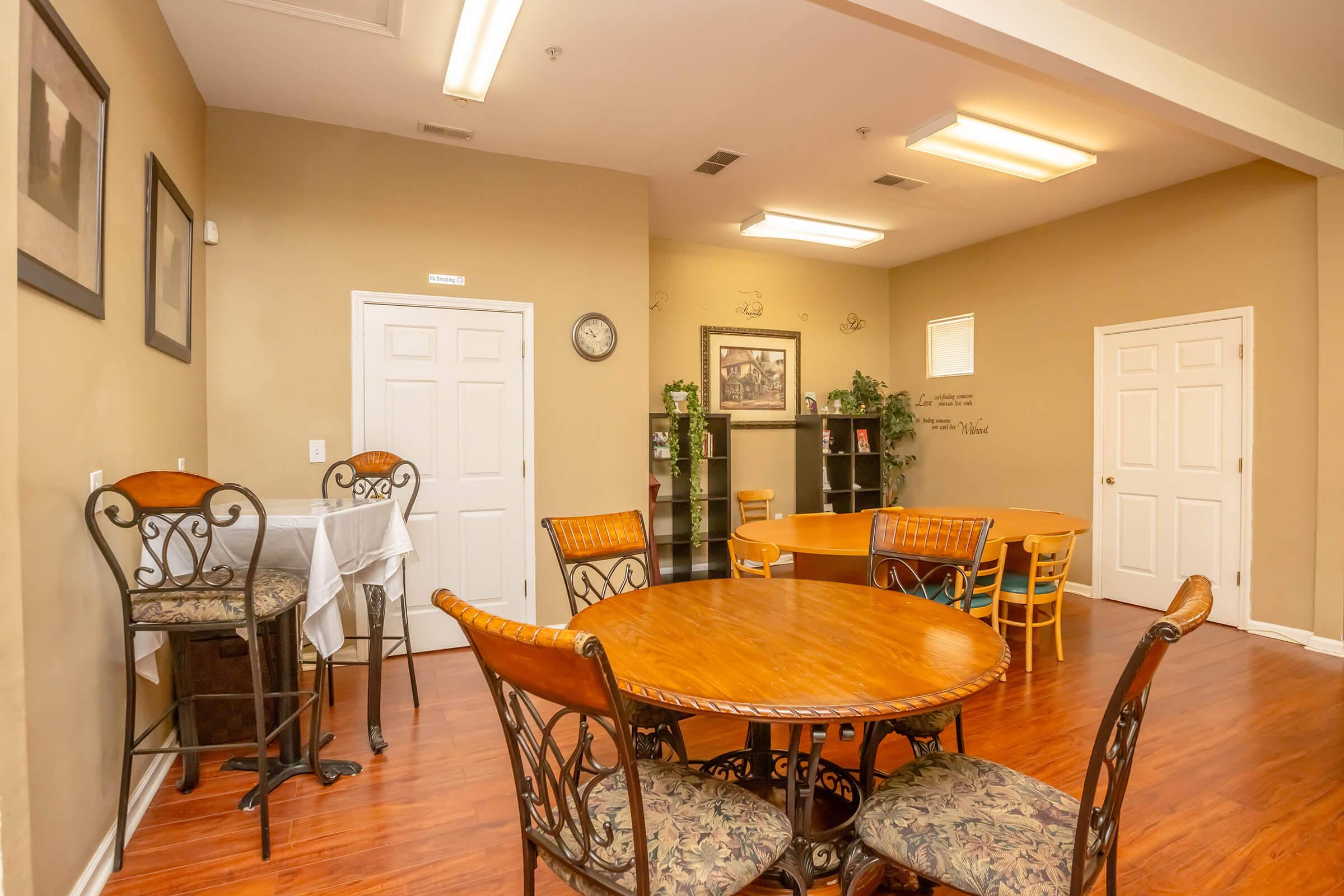A cozy dining area featuring two round wooden tables, one with four chairs and the other with a high-top bar style. The walls are painted in warm tones, with artwork and plants for decoration. A clock and shelves are visible, creating a welcoming atmosphere.