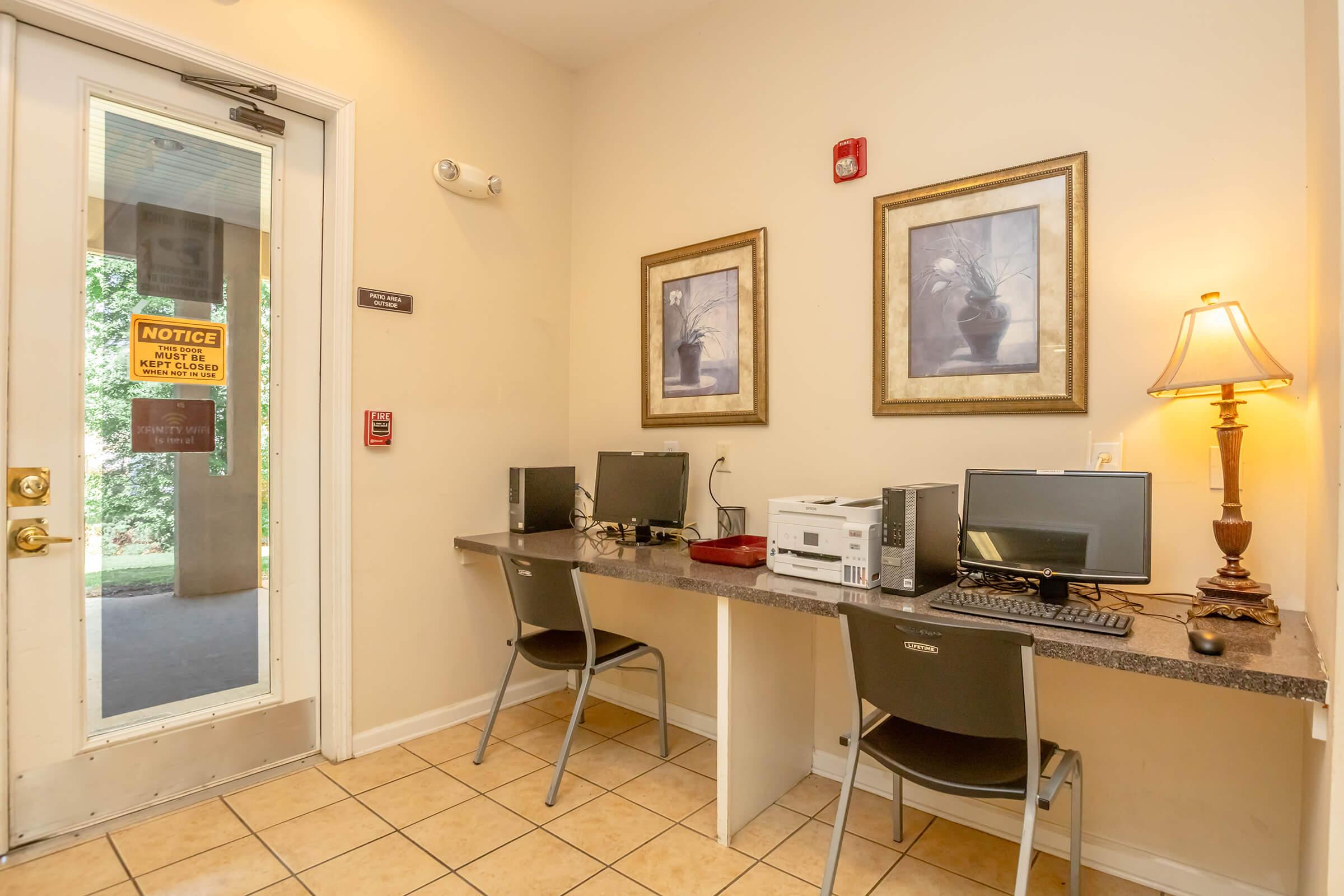 Two workstations with desktop computers are set against a cream-colored wall. Each workstation has a chair and a small lamp on the side. Two framed pictures hang above the desks. A glass door with a notice sign leads outside, providing natural light to the space. The floor is tiled in a light color.