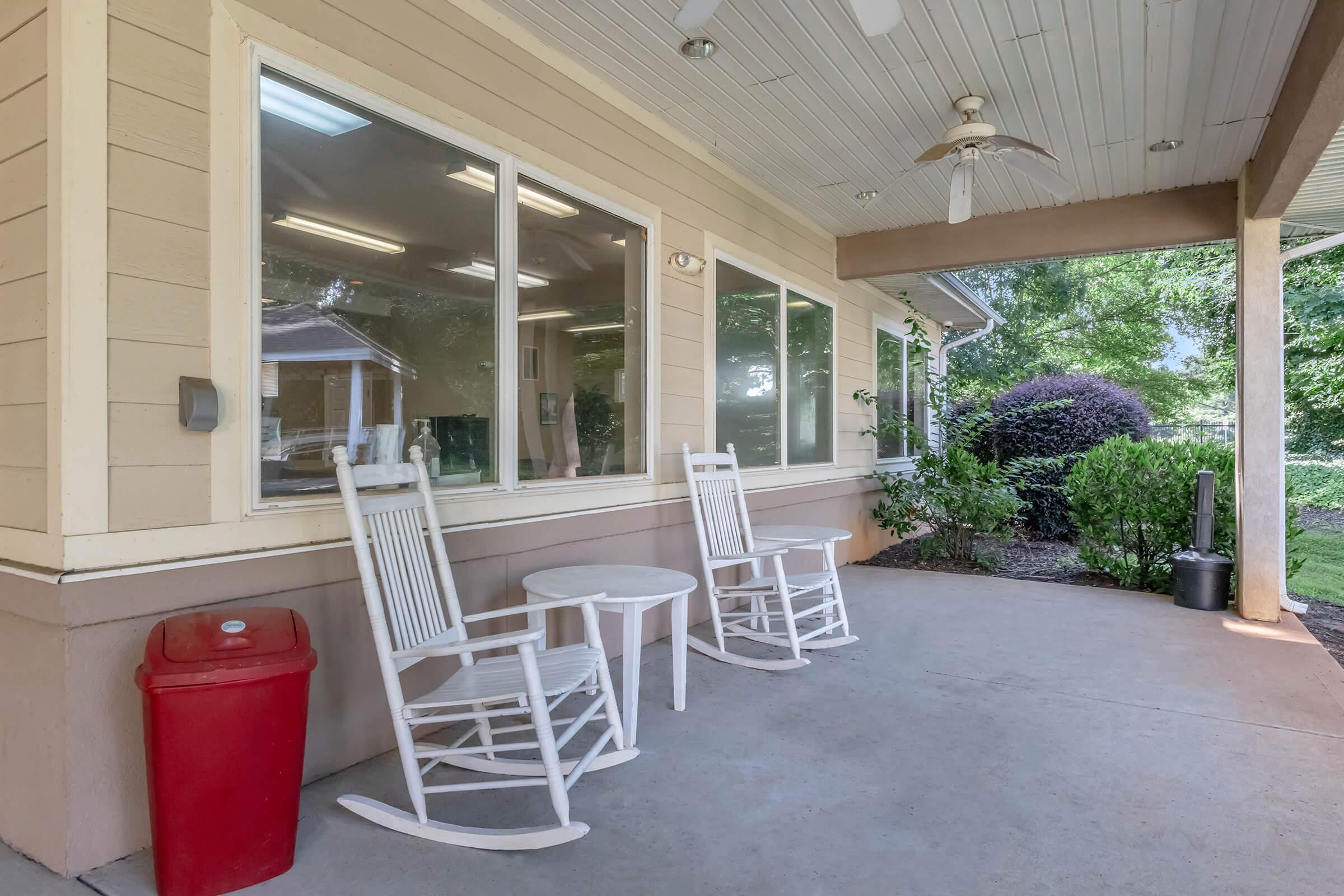 A cozy porch featuring two white rocking chairs and a small table, set against a backdrop of greenery. A red trash can is positioned nearby, and large windows allow natural light to fill the space. The ceiling has a fan, enhancing the inviting atmosphere.