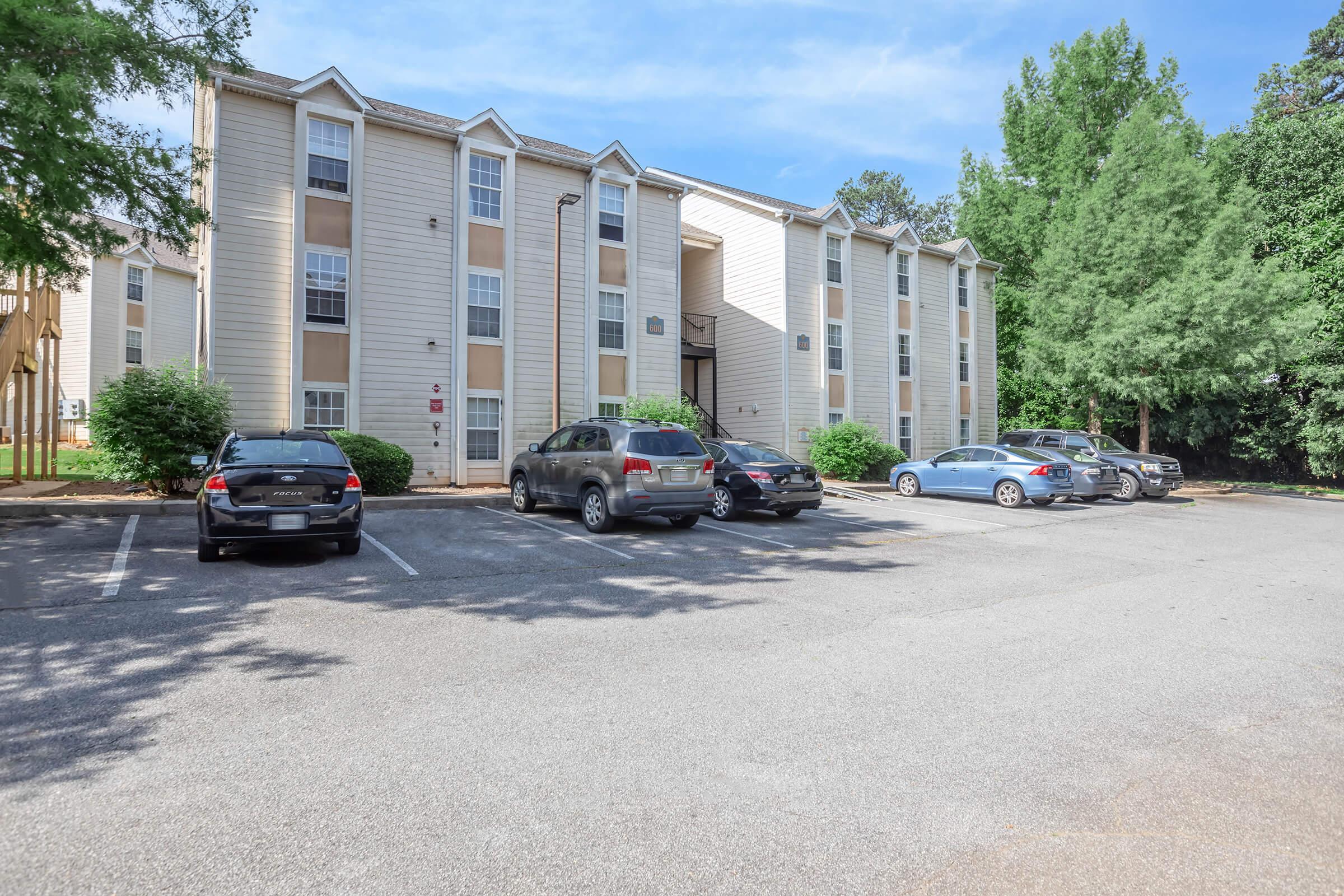 Three-story residential apartment building with a light-colored exterior, surrounded by trees. A parking lot in front has several cars parked, with some spaces available. The sky is clear and blue, suggesting a bright, sunny day.