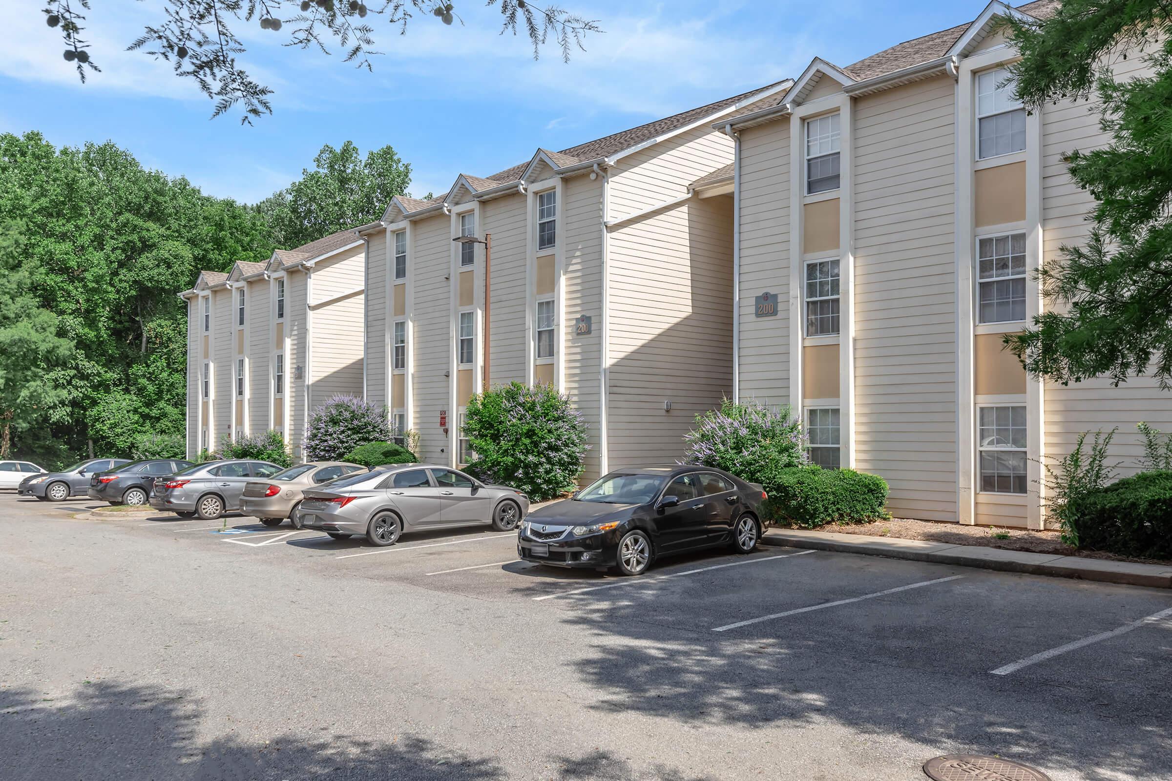 A three-story apartment building with light-colored siding, surrounded by greenery. Several parked cars are visible in the foreground, with a clear blue sky above. The building has multiple windows and a simple architectural design, indicating a residential area.