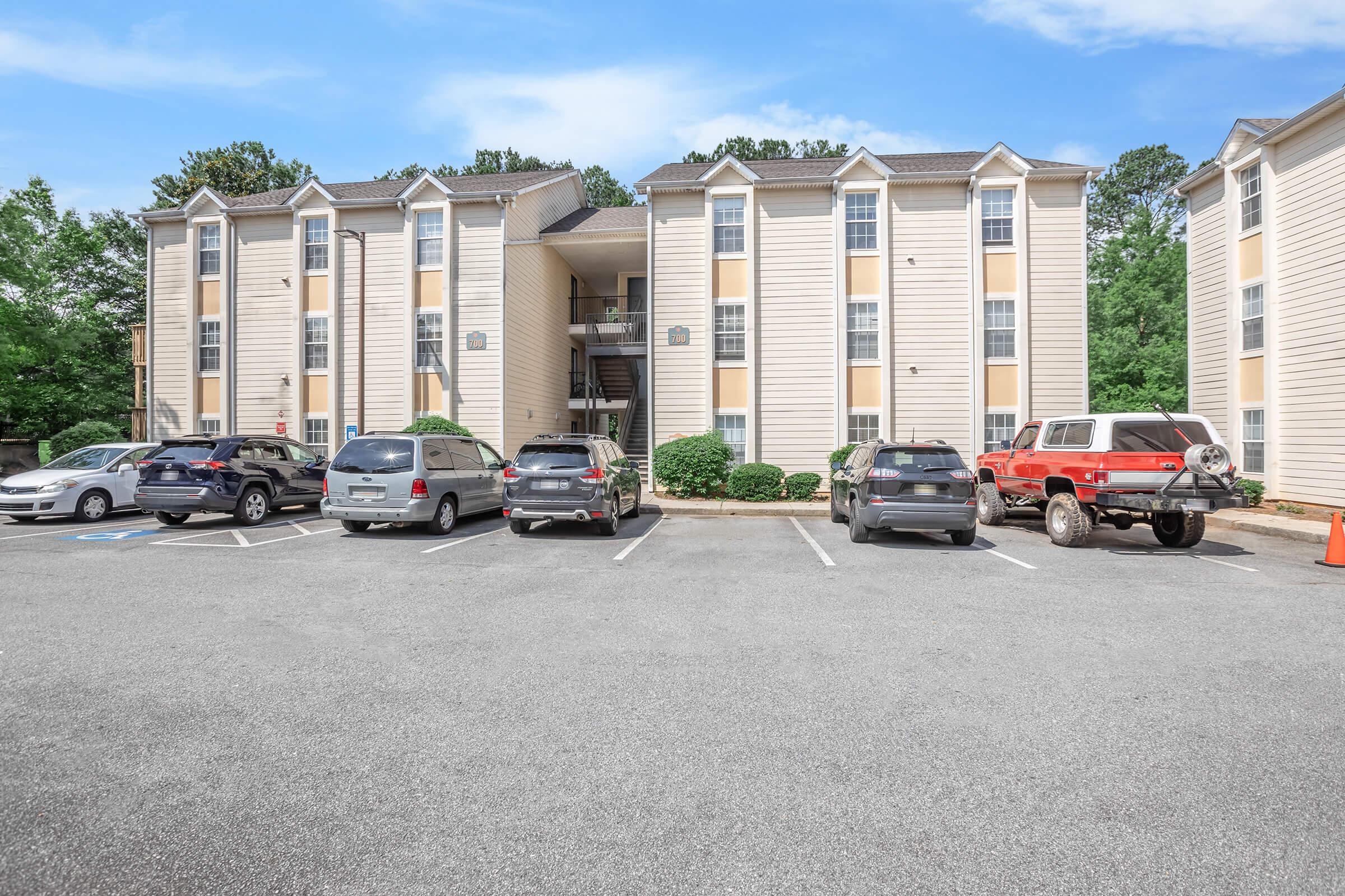 Apartment building with a three-story structure surrounded by a parking lot. Several cars are parked in front, including a red vehicle. The building has a light-colored exterior with multiple windows and balconies. Green trees and blue sky are visible in the background.