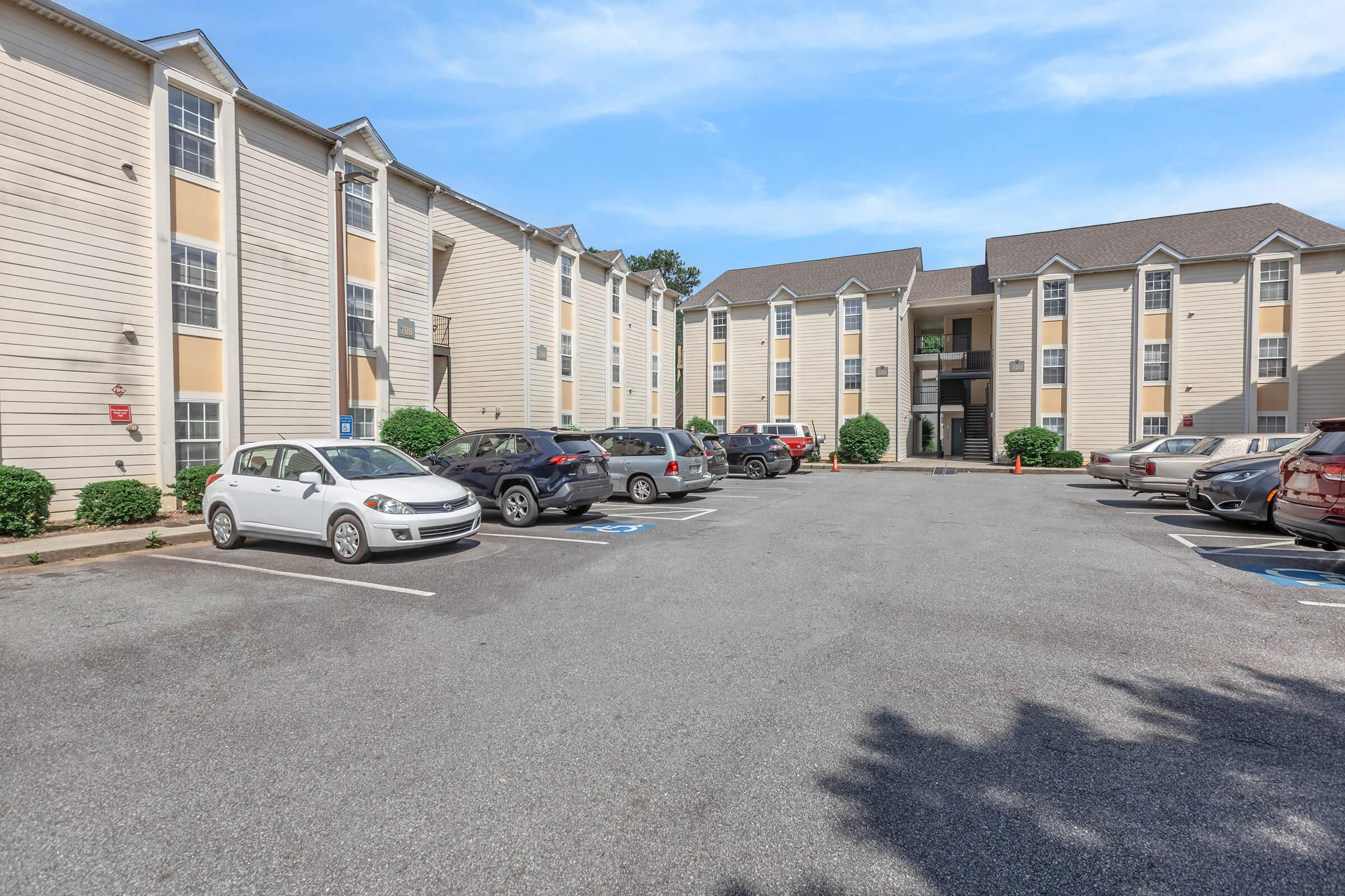 View of an apartment complex featuring two multi-story buildings with light-colored exteriors. The foreground displays a parking lot with multiple cars and landscaped areas. The sky is clear and blue, creating a pleasant atmosphere.