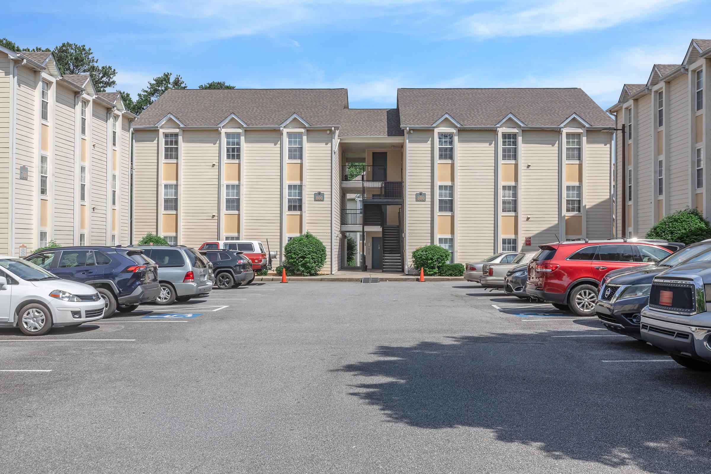A view of a parking lot in front of a multi-story apartment building. Several cars are parked in designated spaces, with a pathway leading up to the entrance of the building. The sky is blue with a few clouds, and there are some green shrubs around the parking area.