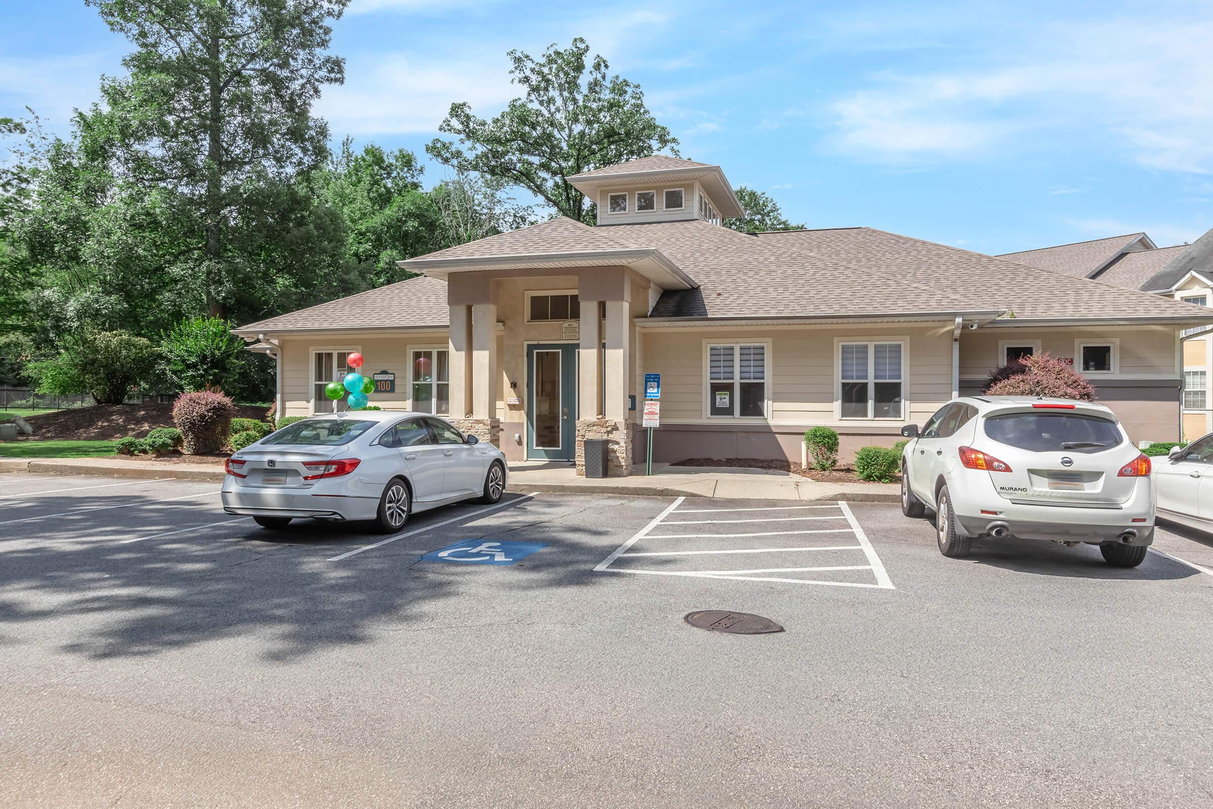 A single-story building with a peaked roof, featuring light-colored siding and a welcoming entrance. Two parked cars, including a white sedan and a silver crossover, are visible in the parking lot, which includes a designated accessible parking space. Colorful balloons adorn the entrance area, suggesting a festive atmosphere.