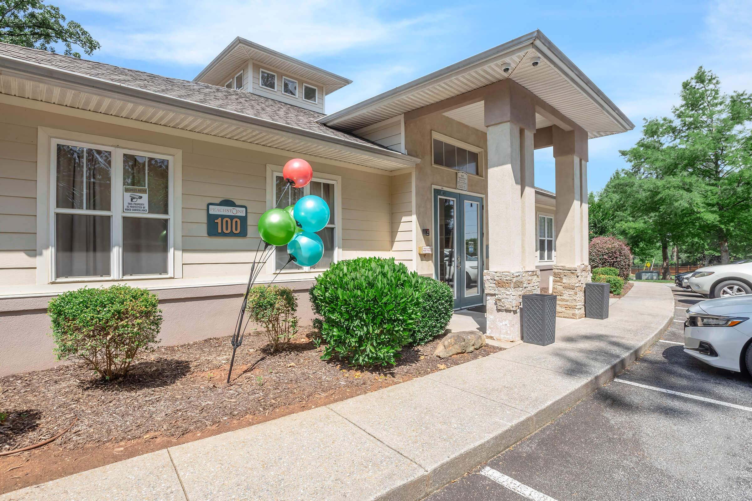 A welcoming exterior view of a building with a light-colored facade, featuring a sign labeled "100." In front, there are colorful balloons in green and red. A well-maintained pathway leads to the entrance, flanked by neatly trimmed bushes, with cars parked nearby and trees in the background.
