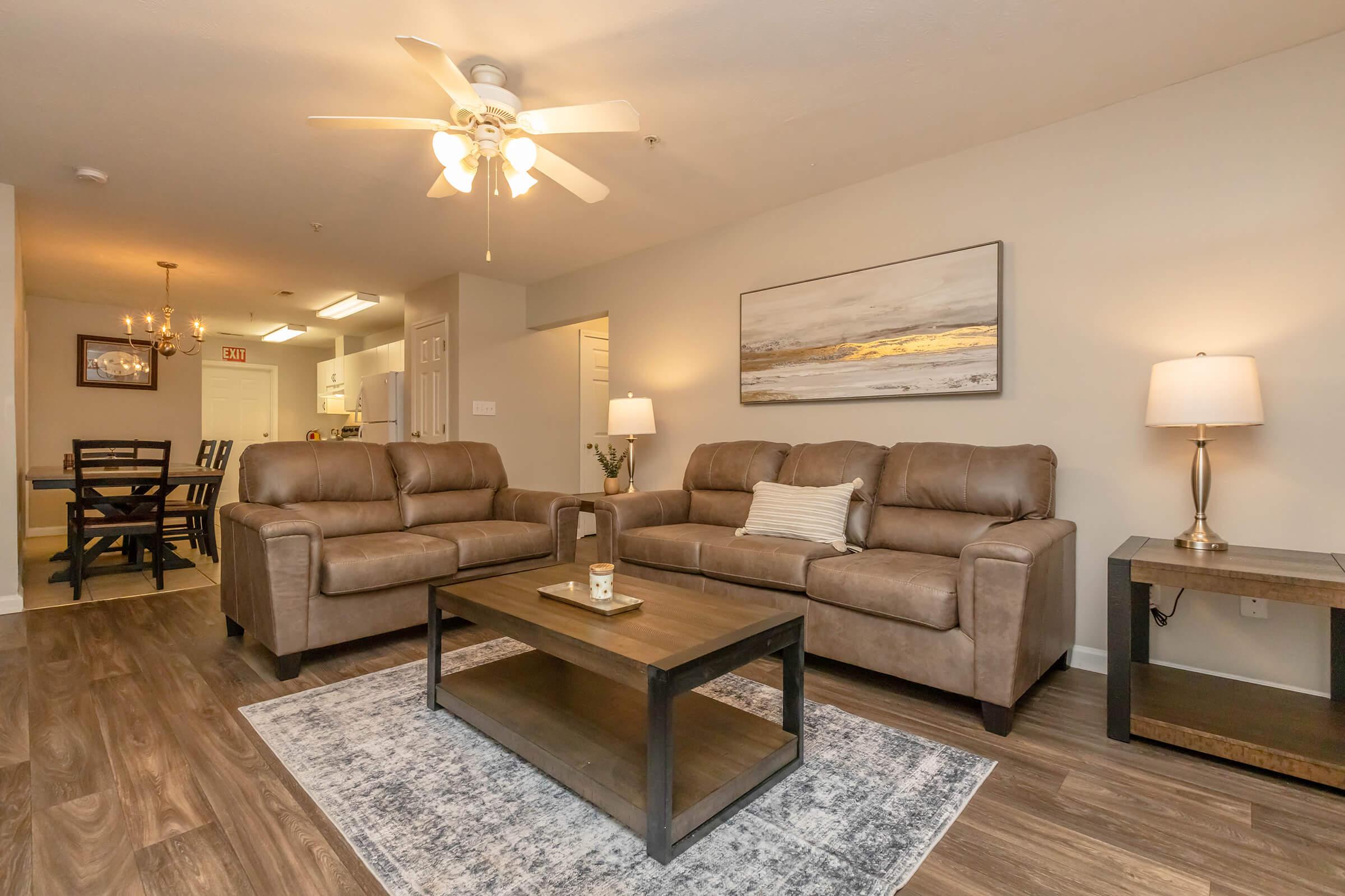 Cozy living room featuring two brown leather sofas, a wooden coffee table with a candle, and a floor lamp. The space has a ceiling fan and is decorated with a wall art piece. In the background, there's a dining area with a table and chairs, enhancing the inviting atmosphere.