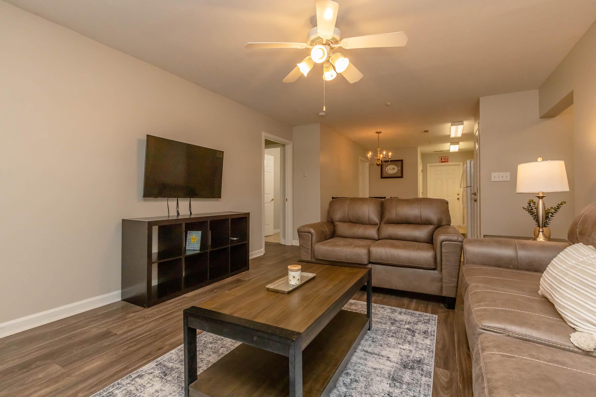 A modern living room featuring a brown leather sofa and loveseat, a dark wooden coffee table, and a flat-screen TV mounted on the wall. The room has warm lighting from a lamp and a ceiling fan, with light-colored walls and wooden flooring.