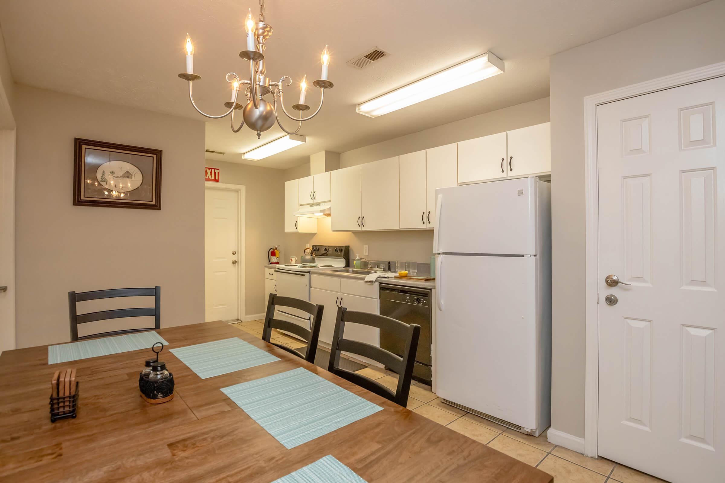 A cozy kitchen featuring white cabinetry, a refrigerator, and a stove. A dining table with wooden chairs is set with light blue placemats. A chandelier hangs above the table, and a wall clock is visible. The room is well-lit, with a door leading outside. Overall, the space is inviting and functional.