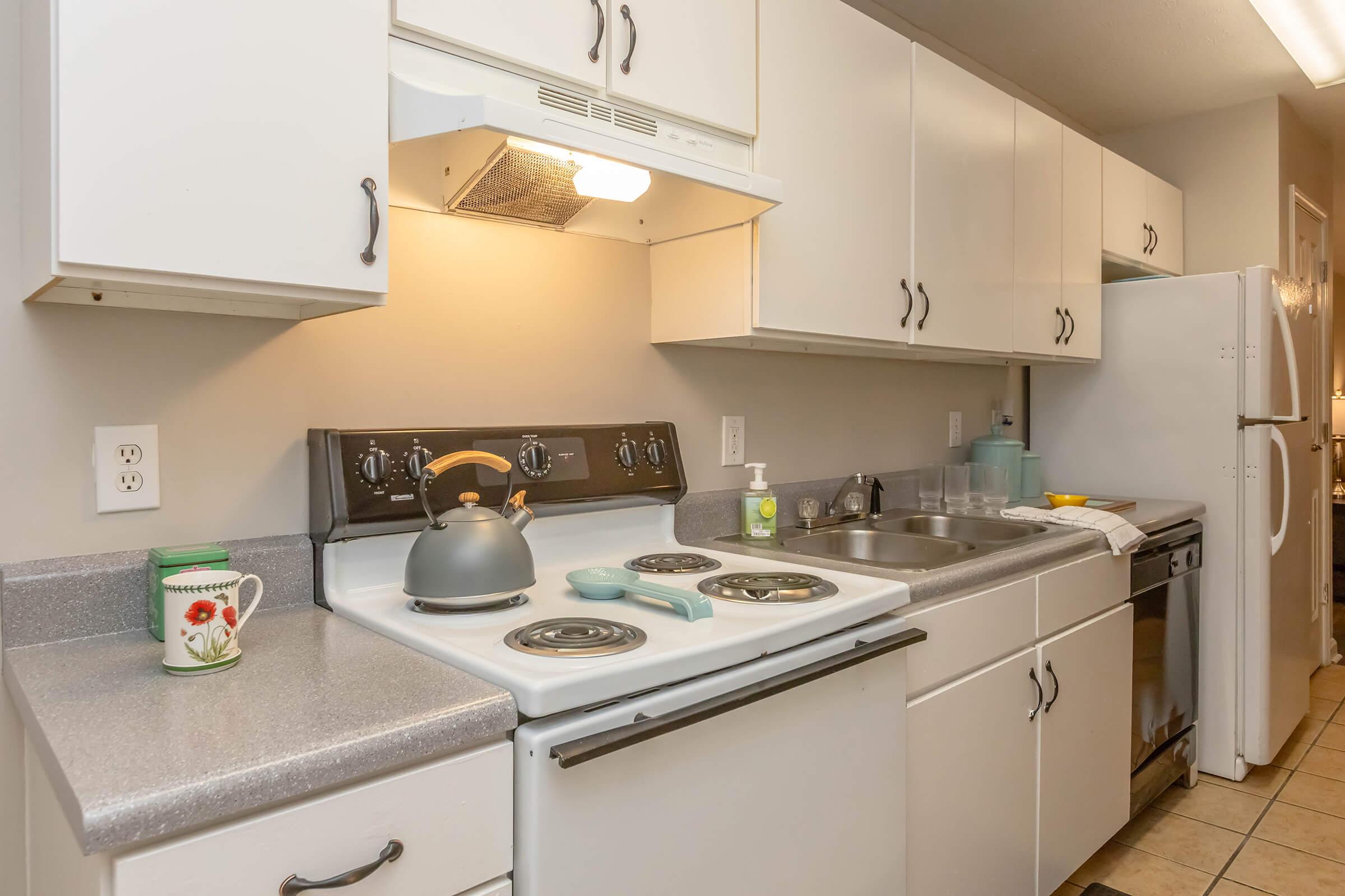 A modern kitchen featuring white cabinets, a gray countertop, a stove with four burners, a kettle on the stove, and various kitchen appliances including a refrigerator and dishwasher. The kitchen is well-lit with a cozy and functional design.