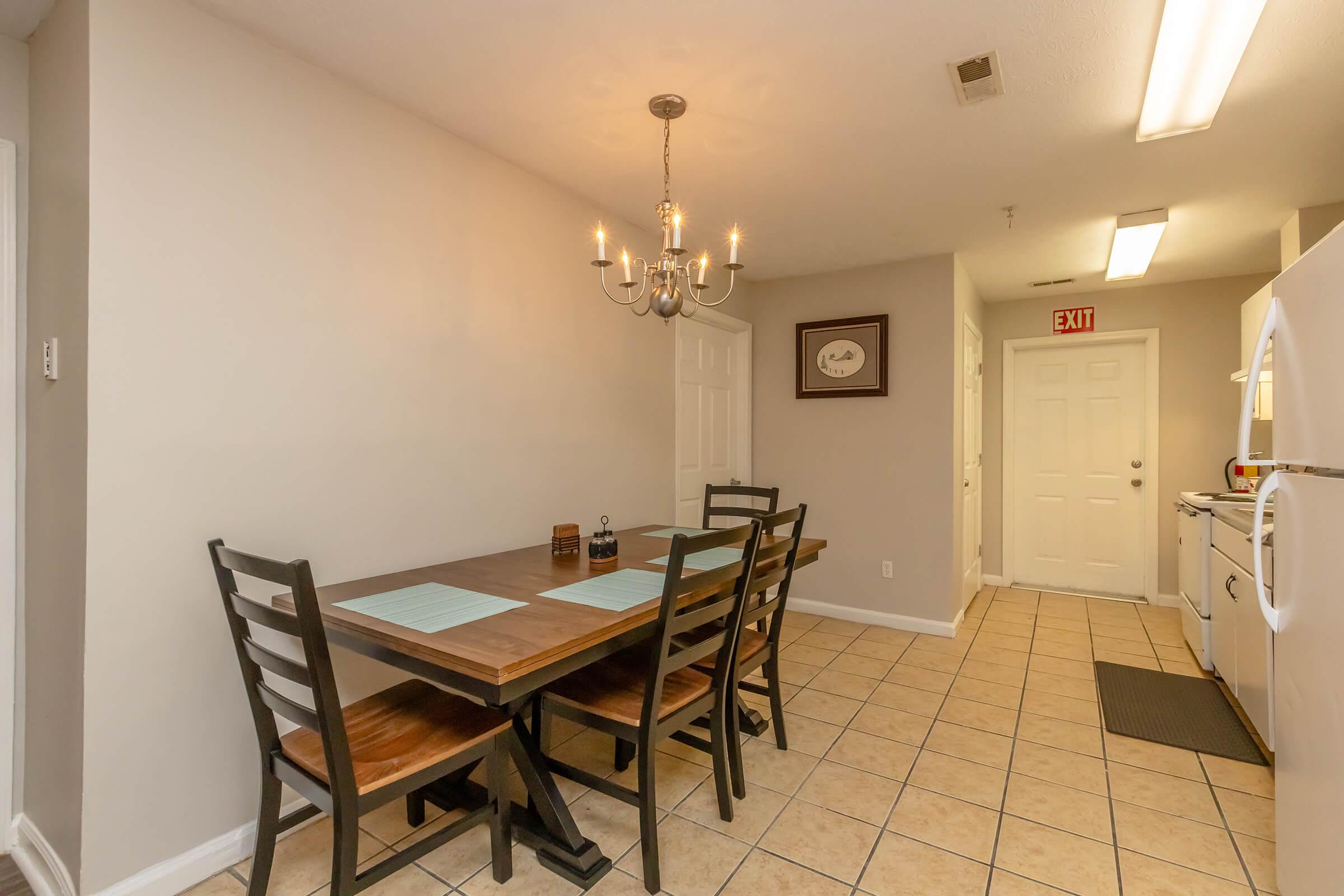 A cozy kitchen featuring a wooden dining table with chairs, adorned with placemats. A chandelier hangs above the table, providing light. The walls are painted in a soft color, and there's a door leading to another room. A white refrigerator and stove are visible at the far end, with tile flooring throughout.