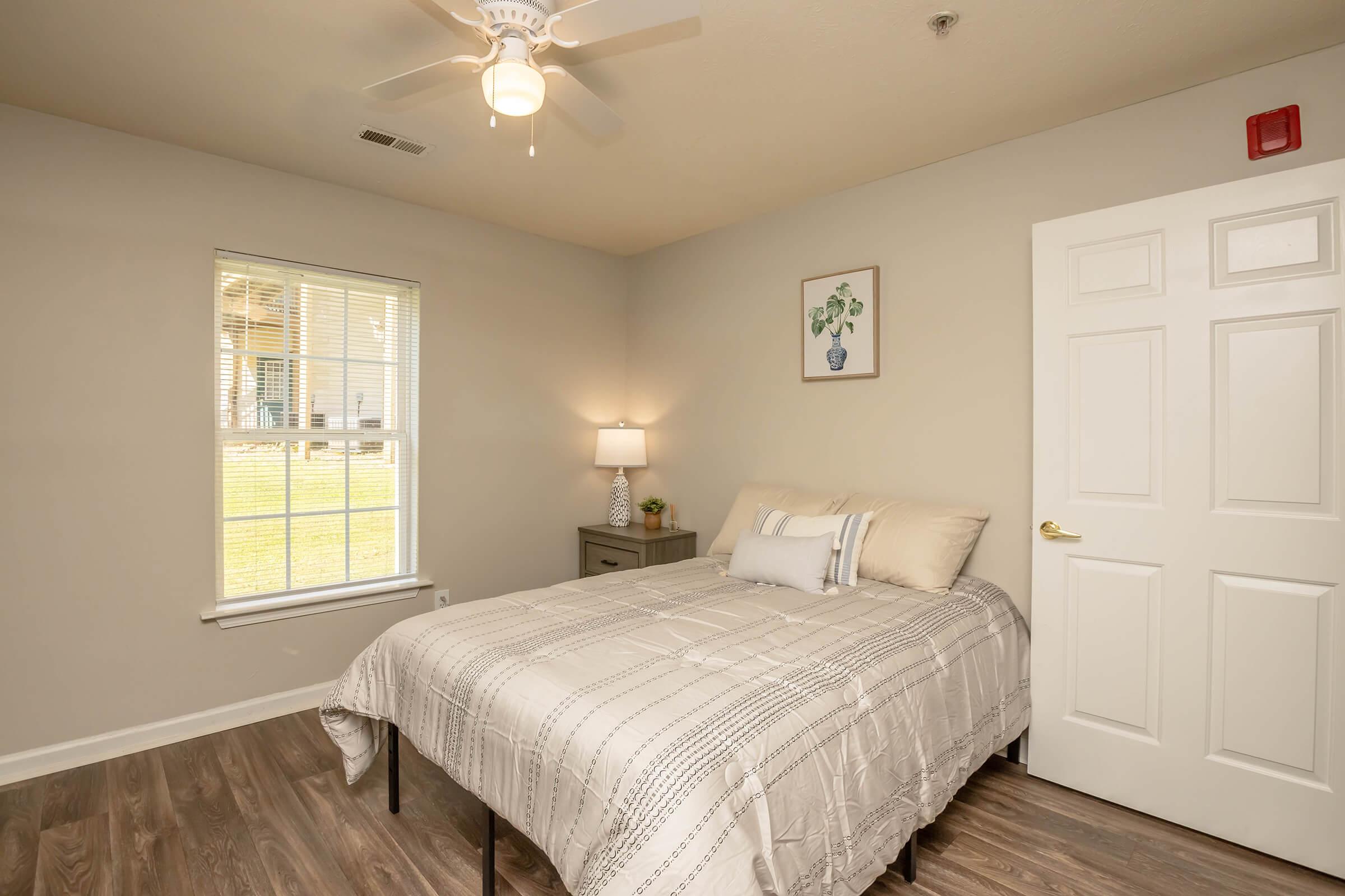 Cozy bedroom featuring a neatly made bed with a light-colored comforter, a bedside table with a lamp, and a framed picture of a plant on the wall. There is a window letting in natural light, showcasing a view of green grass outside, and a closet door on the right. The floor is covered with light wooden planks.