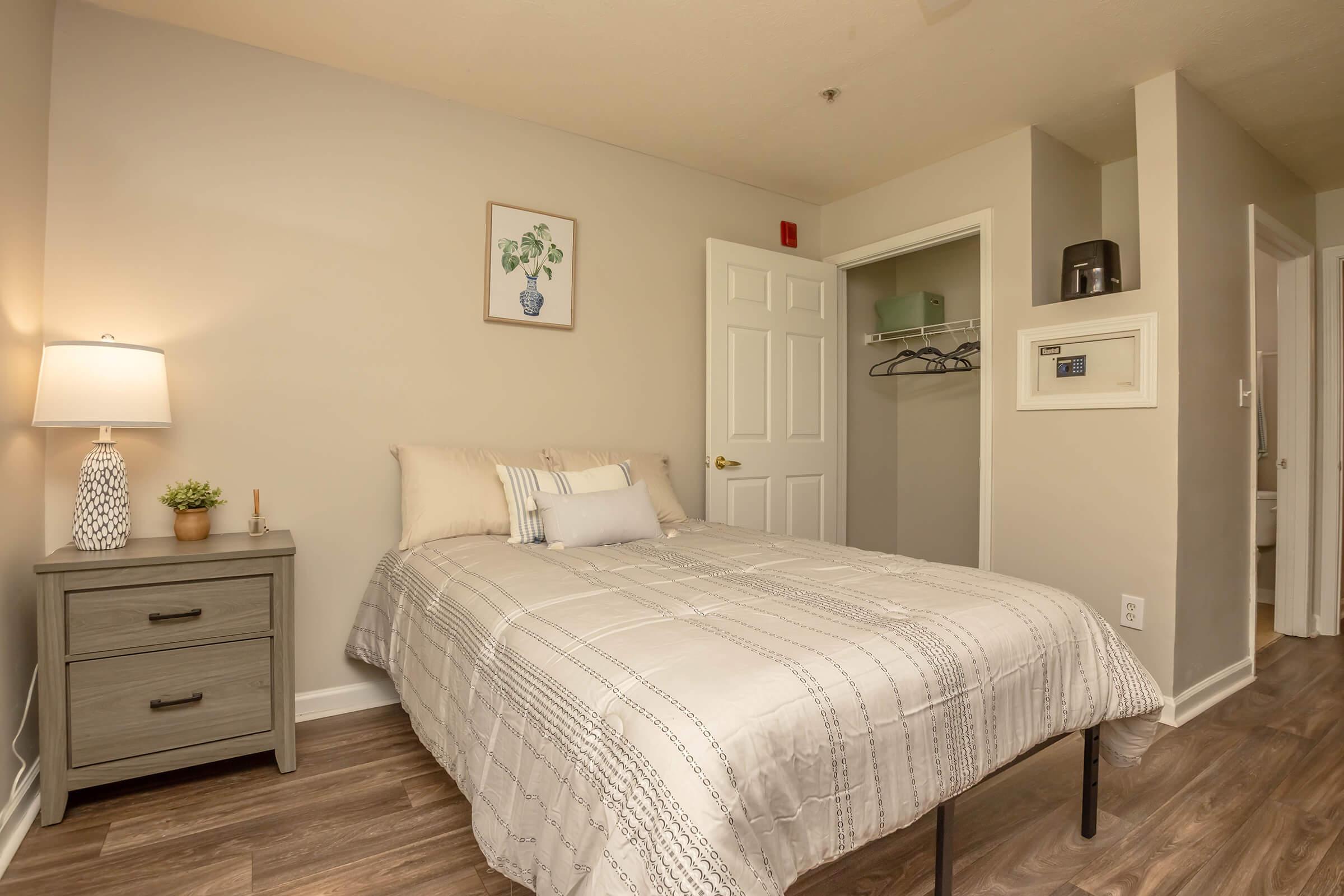 A cozy bedroom featuring a bed with light-colored bedding, a nightstand with a lamp, and a small plant. There's a closet with hanging space visible. The walls are painted in a soft gray tone, and a piece of wall art depicting a plant is displayed above the bed. A bathroom door is slightly visible in the background.