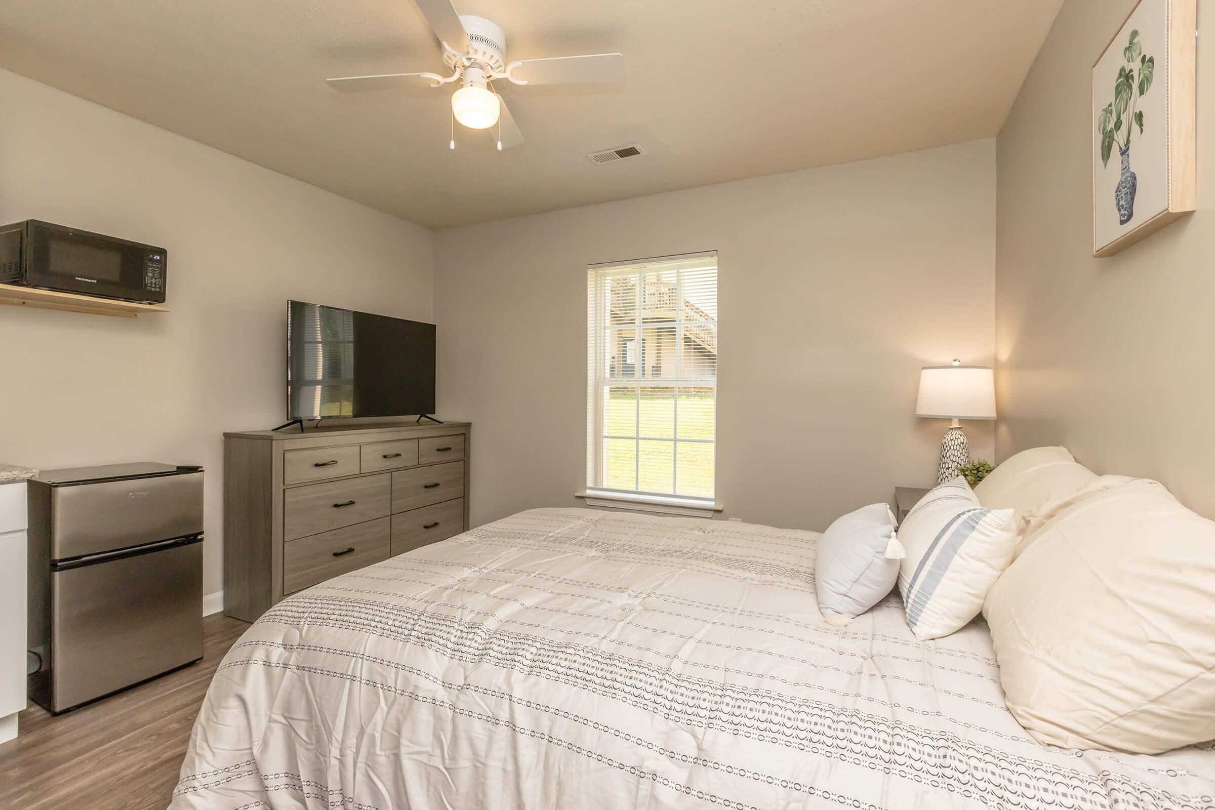 A cozy bedroom featuring a queen-sized bed with white linens, a small dresser, a television on a stand, a microwave, and a mini fridge. Natural light streams in through a window, which overlooks an outdoor area, while a ceiling fan provides ventilation. Decorative elements add a homey touch.