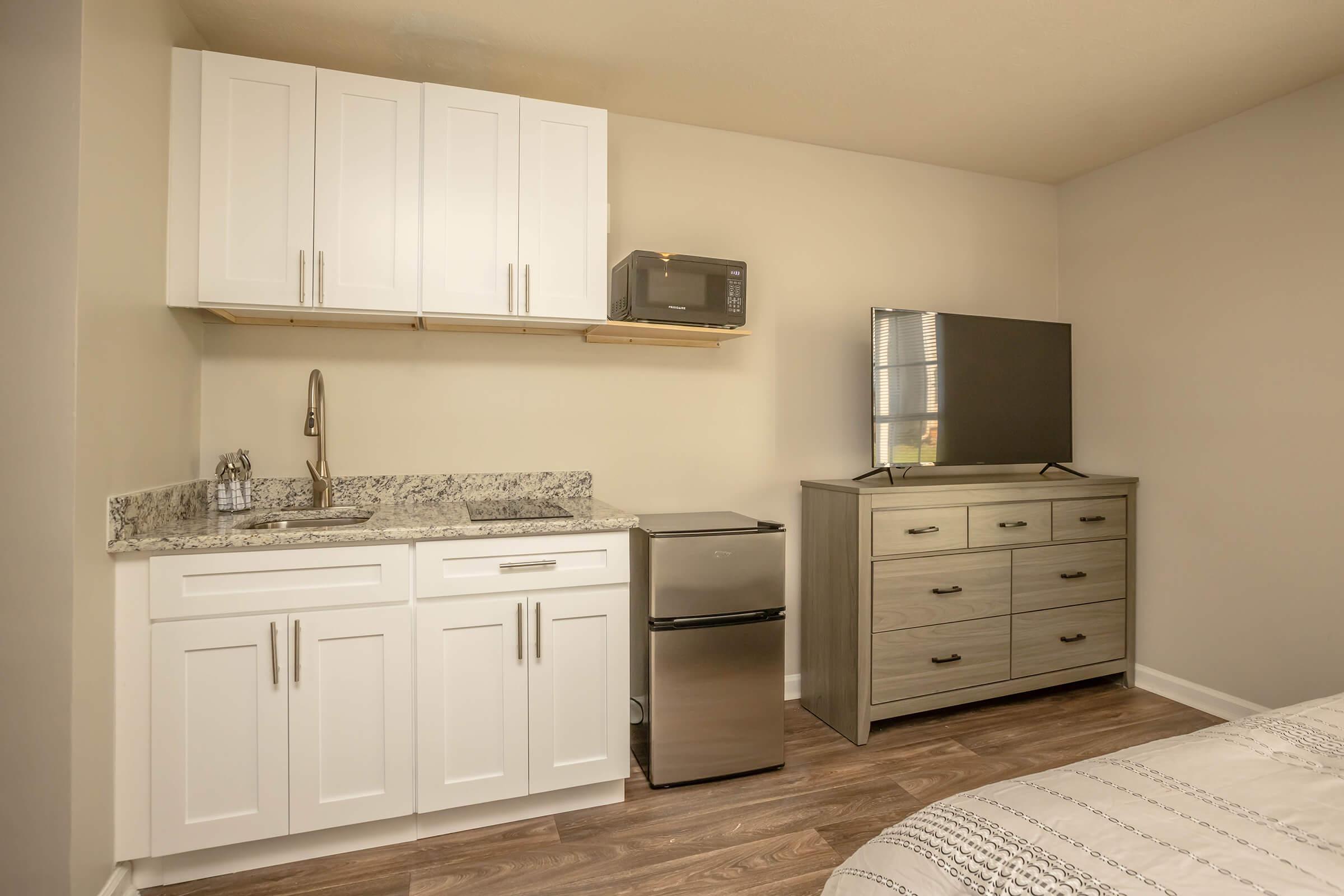 A small living space features a kitchenette with white cabinets, a granite countertop, a sink, a microwave, and a compact refrigerator. There's a television on top of a dresser with multiple drawers, and light-colored walls complement the wooden flooring. The overall design is modern and minimalist.