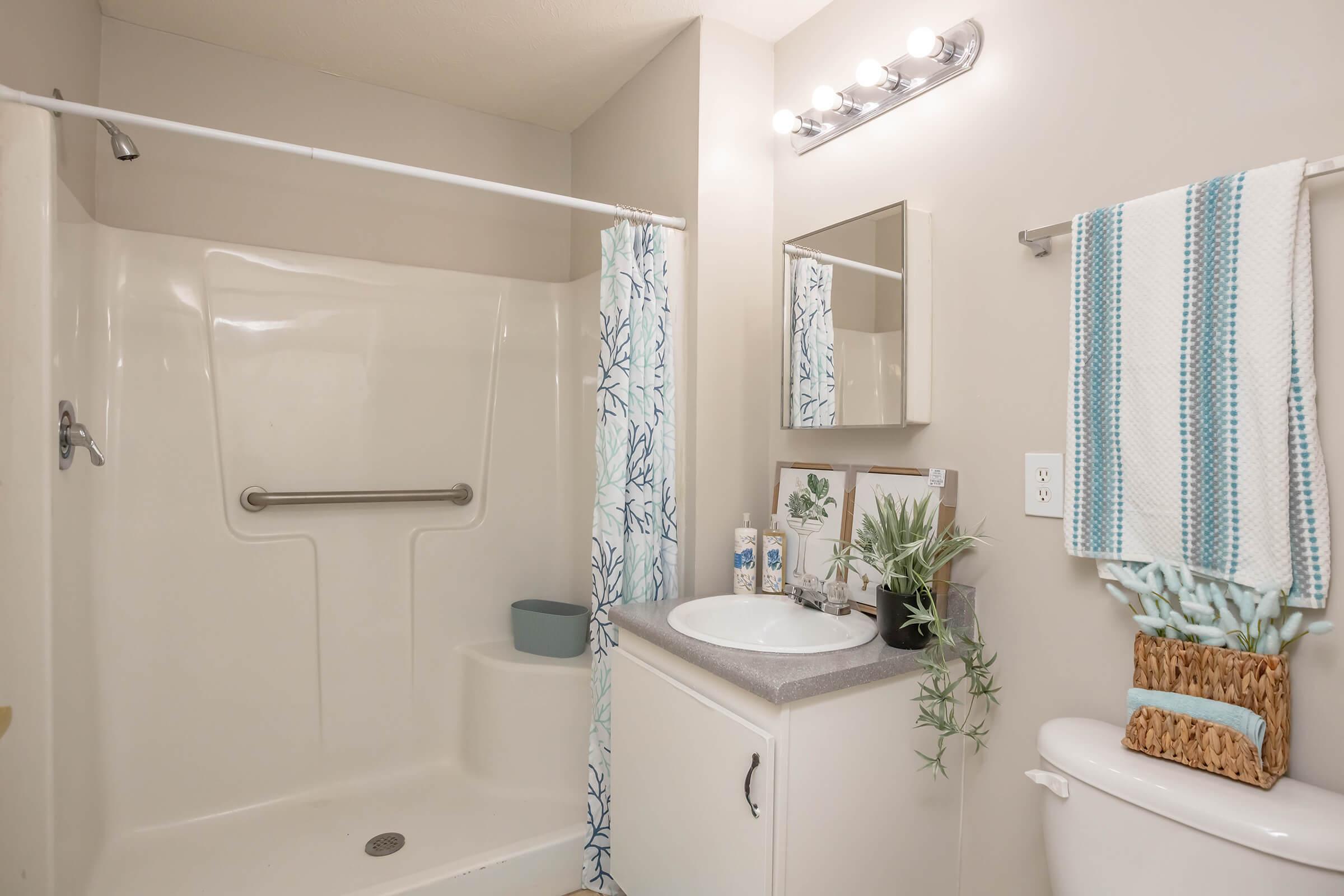 A clean and modern bathroom featuring a shower with a grab bar, a small sink with a vanity mirror, and decorative elements like a towel rack and a basket. The color scheme includes light tones with touches of blue and green, and there are potted plants for a refreshing look.