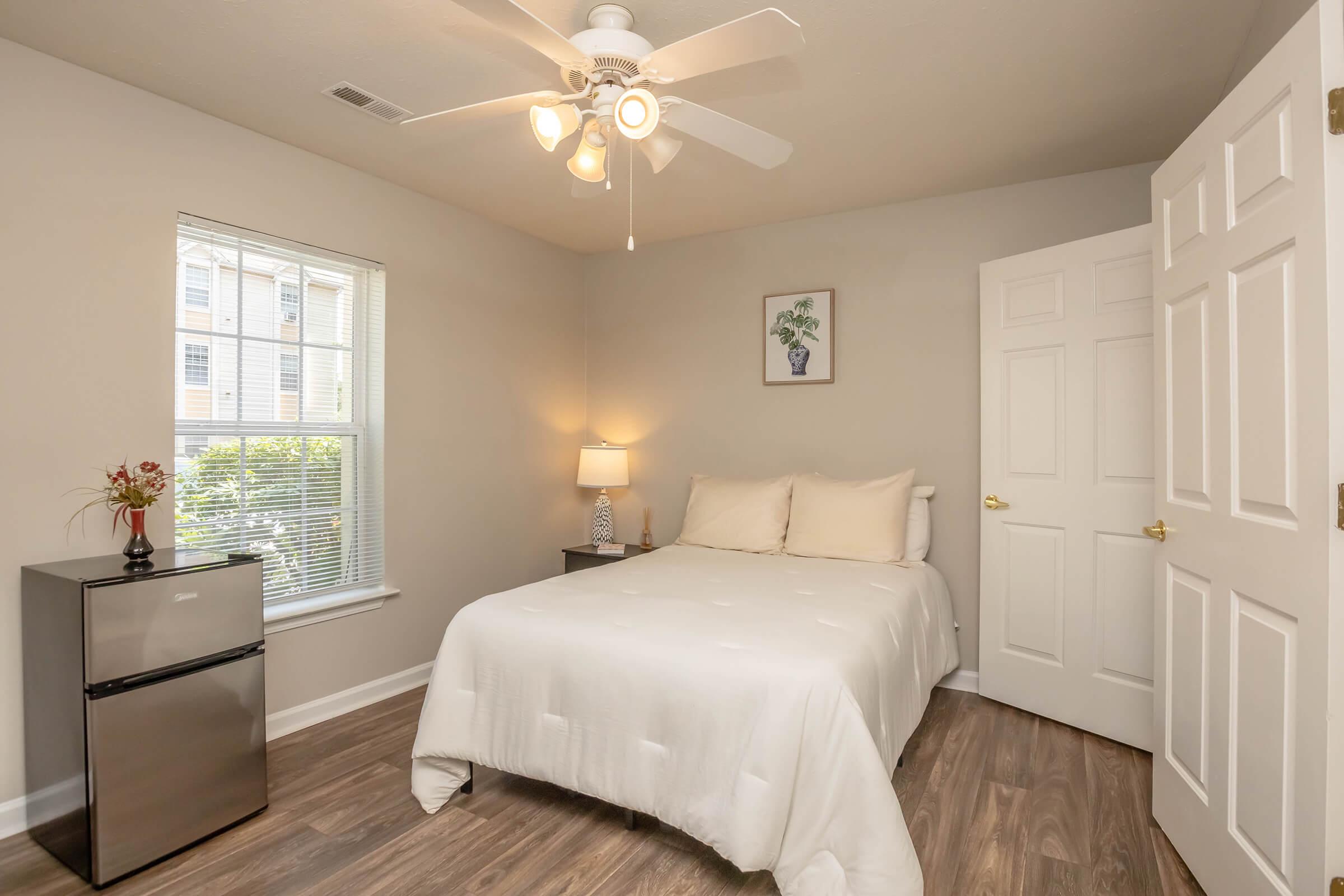 A cozy bedroom featuring a queen-sized bed with white bedding, a bedside lamp, a small refrigerator, and a ceiling fan. The room has a window with blinds allowing natural light, and a decorative plant hangs on the wall. The flooring is wooden and the walls are painted a neutral color.