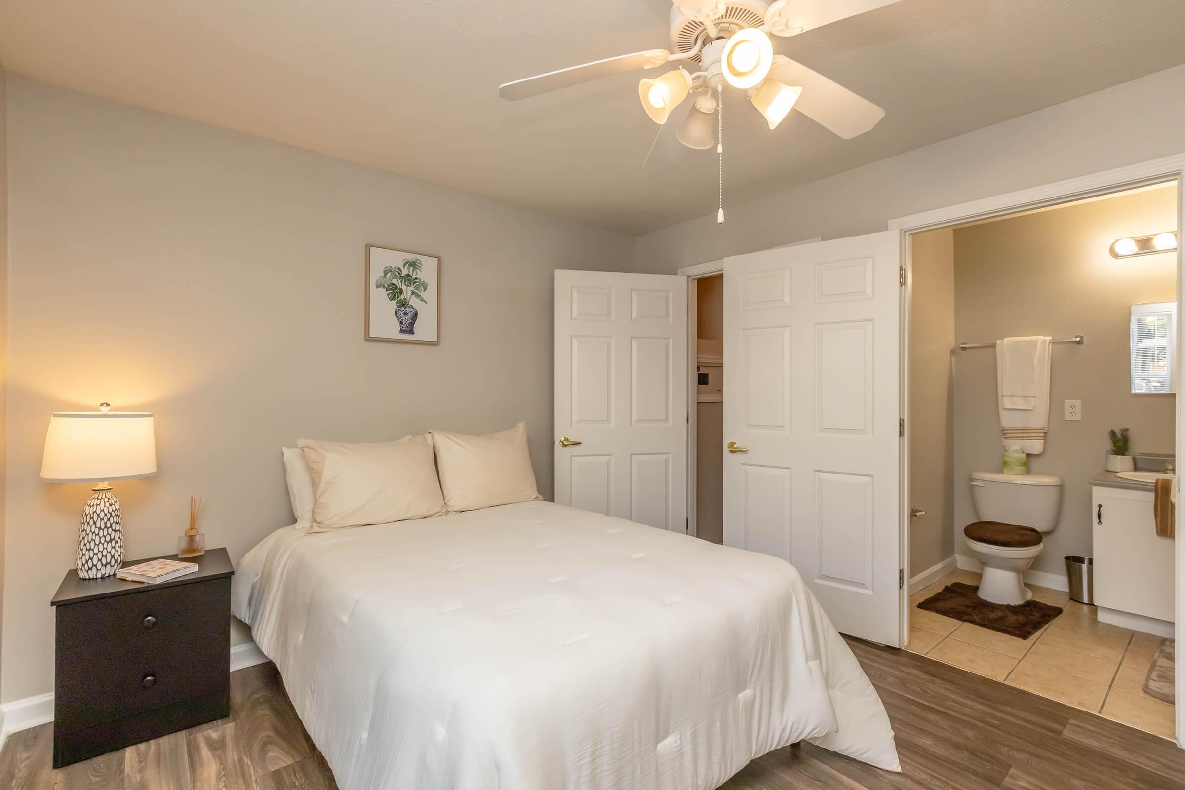 Cozy bedroom featuring a queen-sized bed with light bedding, a bedside lamp on a dark nightstand, and a framed plant picture on the wall. Double doors lead to a bathroom with a bathtub, sink, and toilet, all set in a neutral color scheme with wood-like flooring.