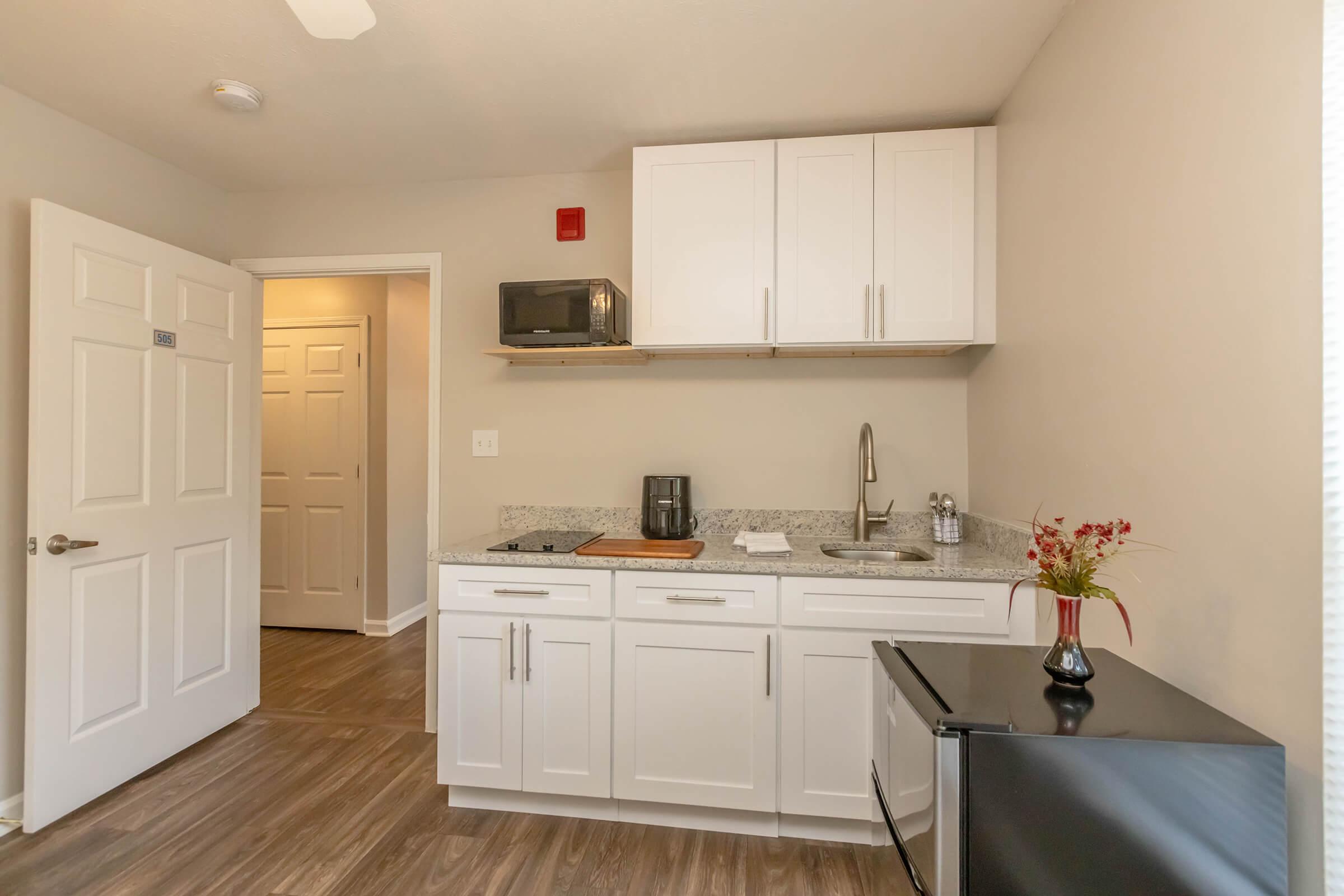 A well-lit kitchenette featuring white cabinets, a granite countertop, a microwave, a coffee maker, and a small black refrigerator. The flooring is wood-like, and there's a small vase with flowers on the counter. A door in the background leads to a hallway.