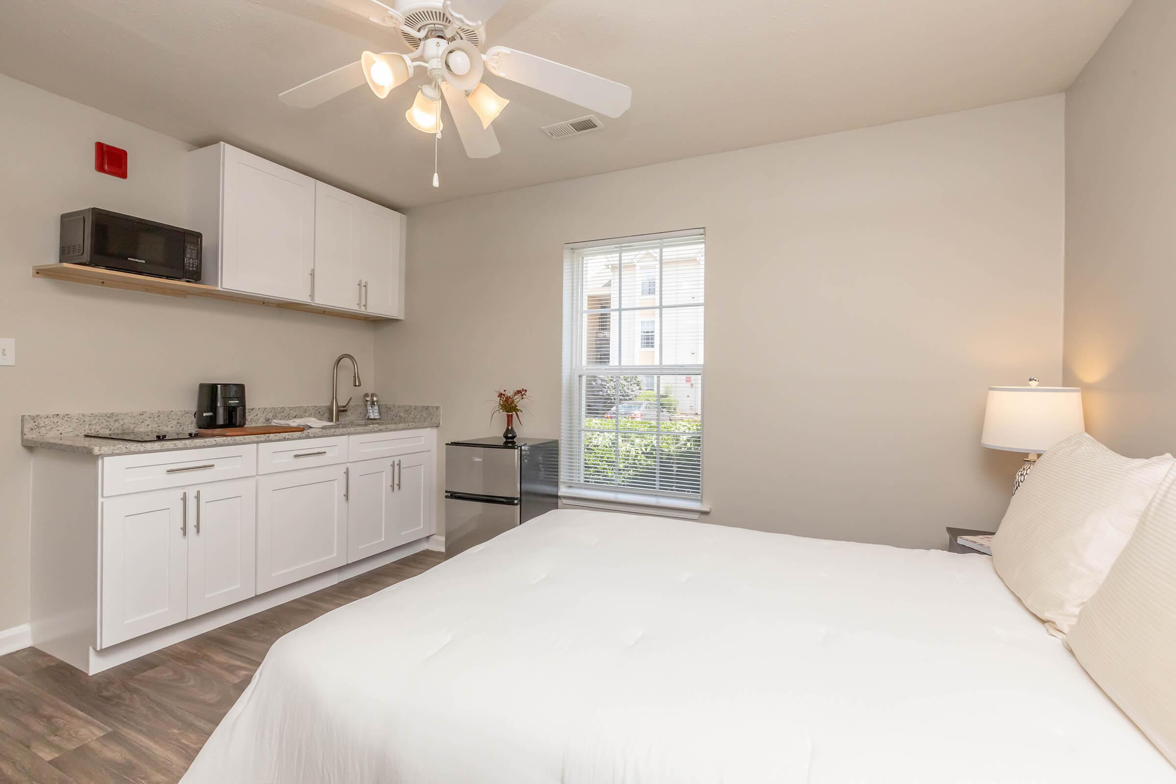 A cozy room featuring a queen-sized bed, a kitchenette with a microwave and sink, and a small fridge. Natural light enters through the window, with simple decor and a lamp providing a warm atmosphere. The space is designed for comfort and functionality.
