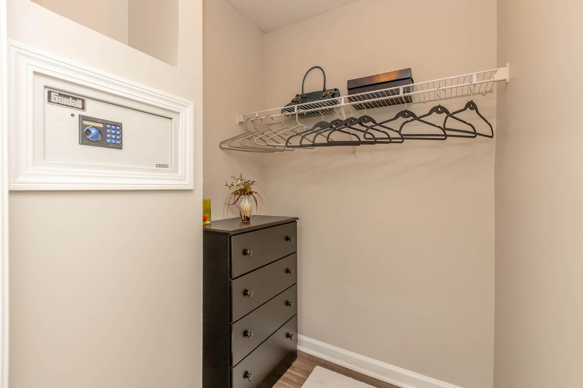 A small closet featuring a wire shelf with several hangers, a dresser with multiple drawers, and a wall-mounted safe. The walls are painted in a light color, creating a clean and organized space.