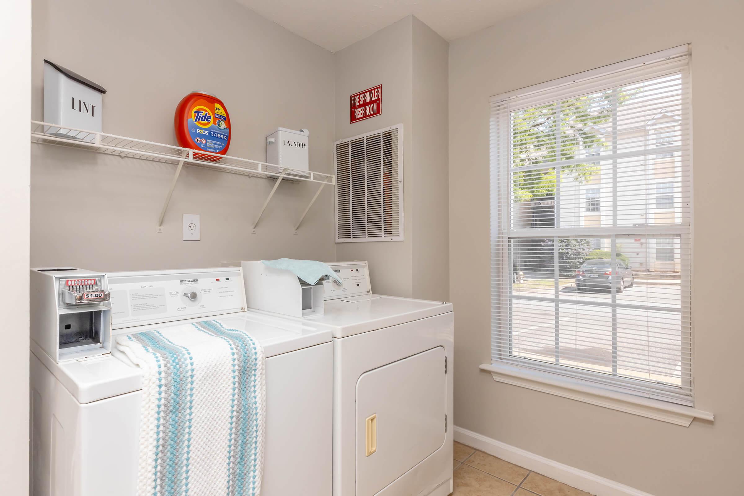 A laundry room featuring two washers and a dryer. The appliances are against a light gray wall, with a shelf above holding a bottle of detergent. A blue and white towel is draped over the dryer. A window provides natural light, showing a view of a parking area outside. A "Lint" sign is visible on the wall.