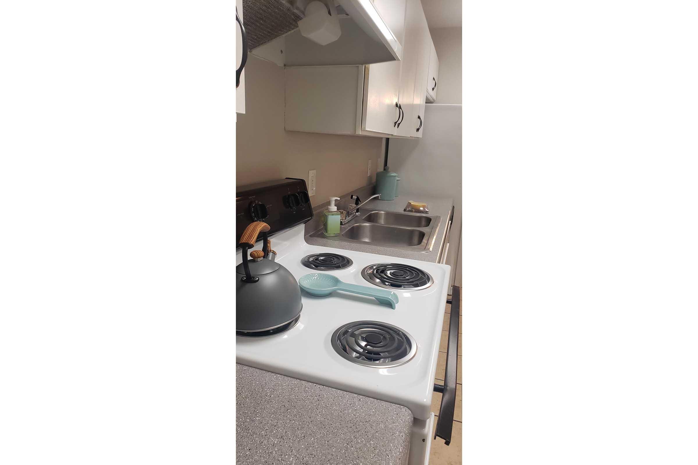 A modern kitchen featuring a white stove with black burners, a kettle on the stove, and a green slotted spoon. A stainless steel sink is visible with a bottle of soap nearby. The cabinets are white, and the overall color scheme is light and neutral.