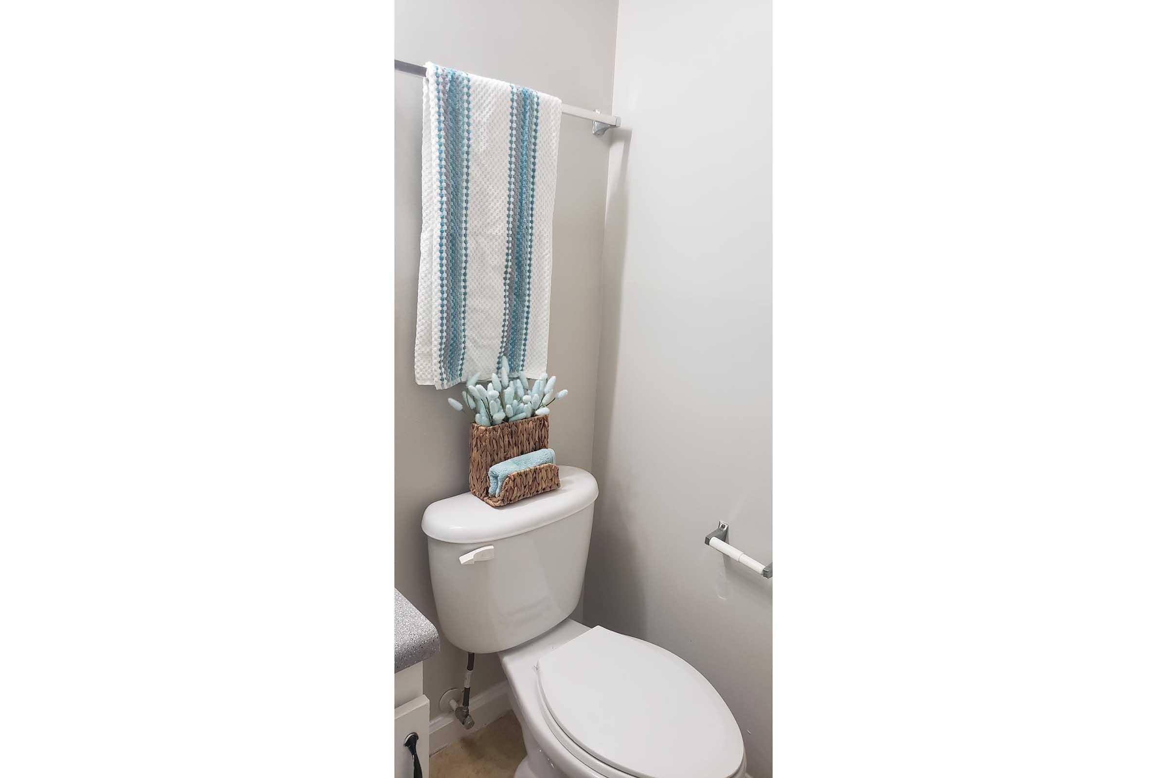 A clean bathroom scene featuring a white toilet with a woven basket on top, holding rolled towels. A striped blue and white towel hangs on a bar on the wall, and there's a small countertop with a light-colored surface. The walls are painted in a soft neutral color.