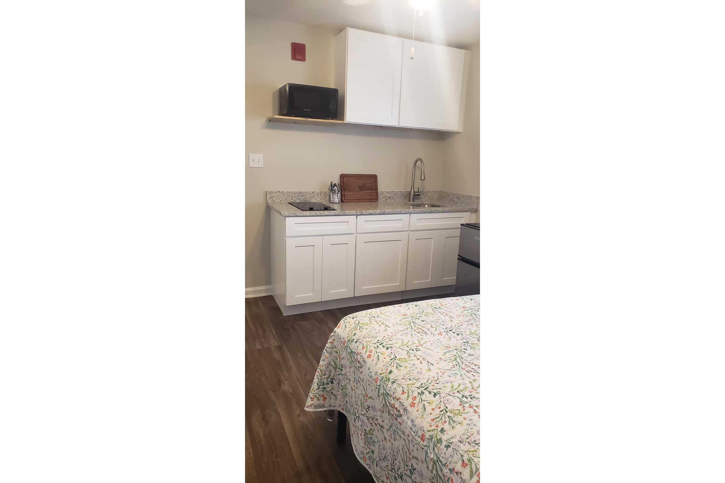 A compact kitchen area featuring white cabinetry, a granite countertop, a sink, and a microwave on a shelf above. The kitchen is located next to a bed with a floral-patterned bedspread. The flooring is dark laminate, and the walls are painted a light color.