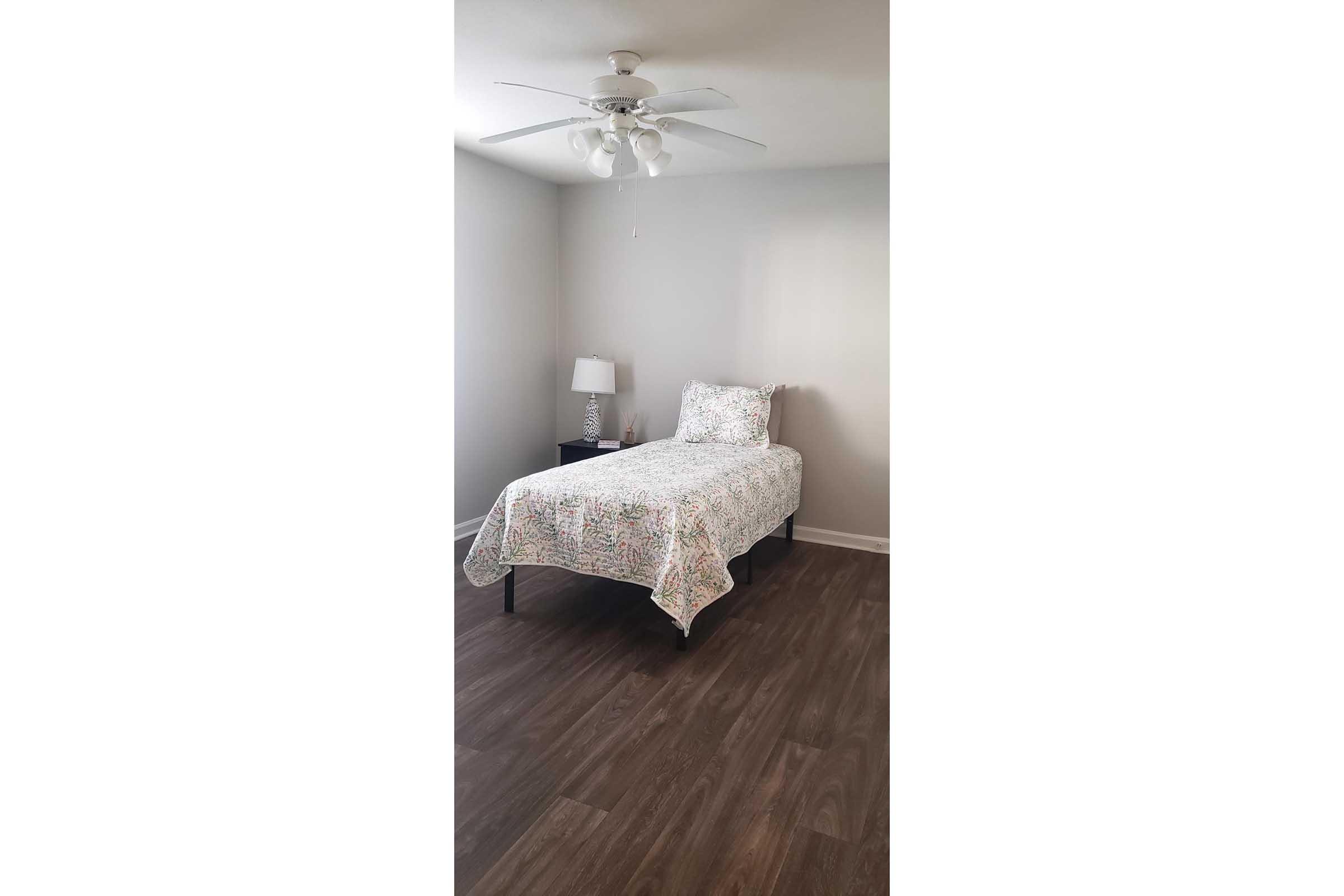 A simple bedroom featuring a single bed covered with a floral quilt, a matching bedside lamp on a nightstand, and a ceiling fan. The walls are painted light gray, and the floor has a dark wooden finish, creating a clean and minimalist aesthetic.