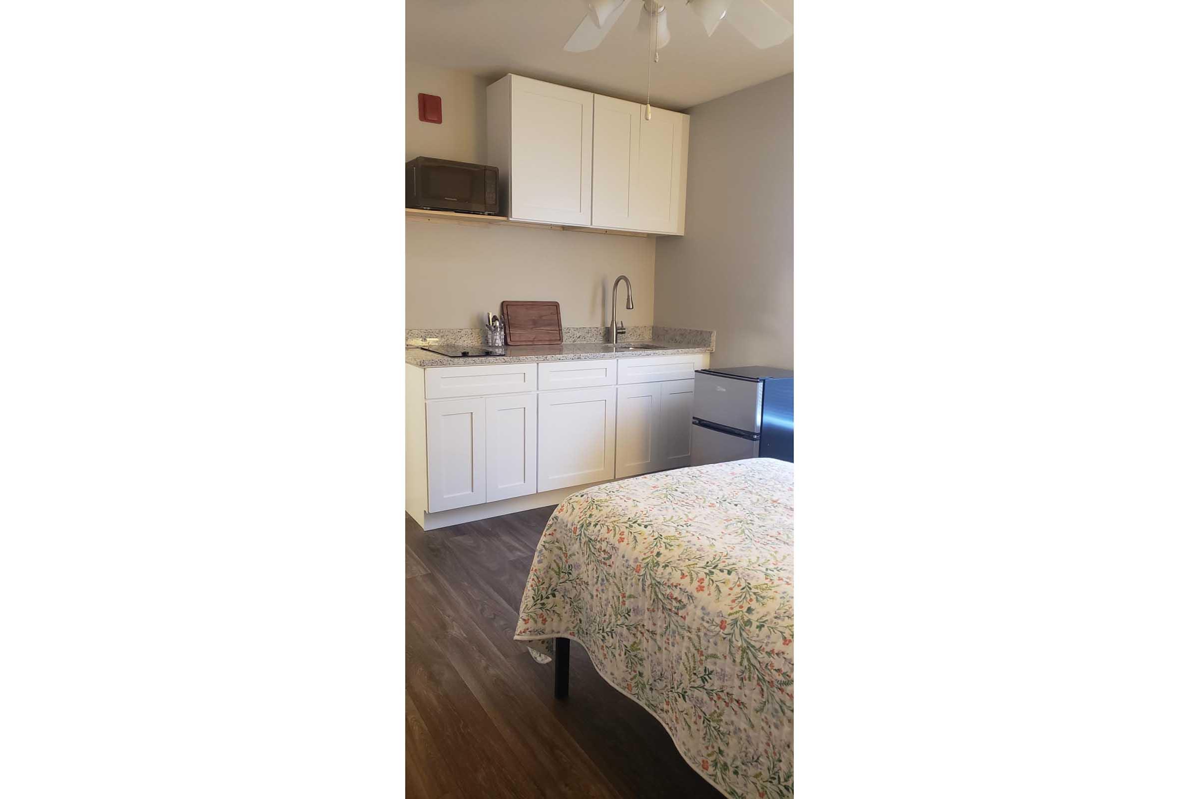 A small kitchen area featuring white cabinets and a countertop with a sink. There's a microwave above the counter, and a small refrigerator is visible. In the foreground, a bed is partially visible with a floral-patterned bedspread, set against a neutral-colored wall.