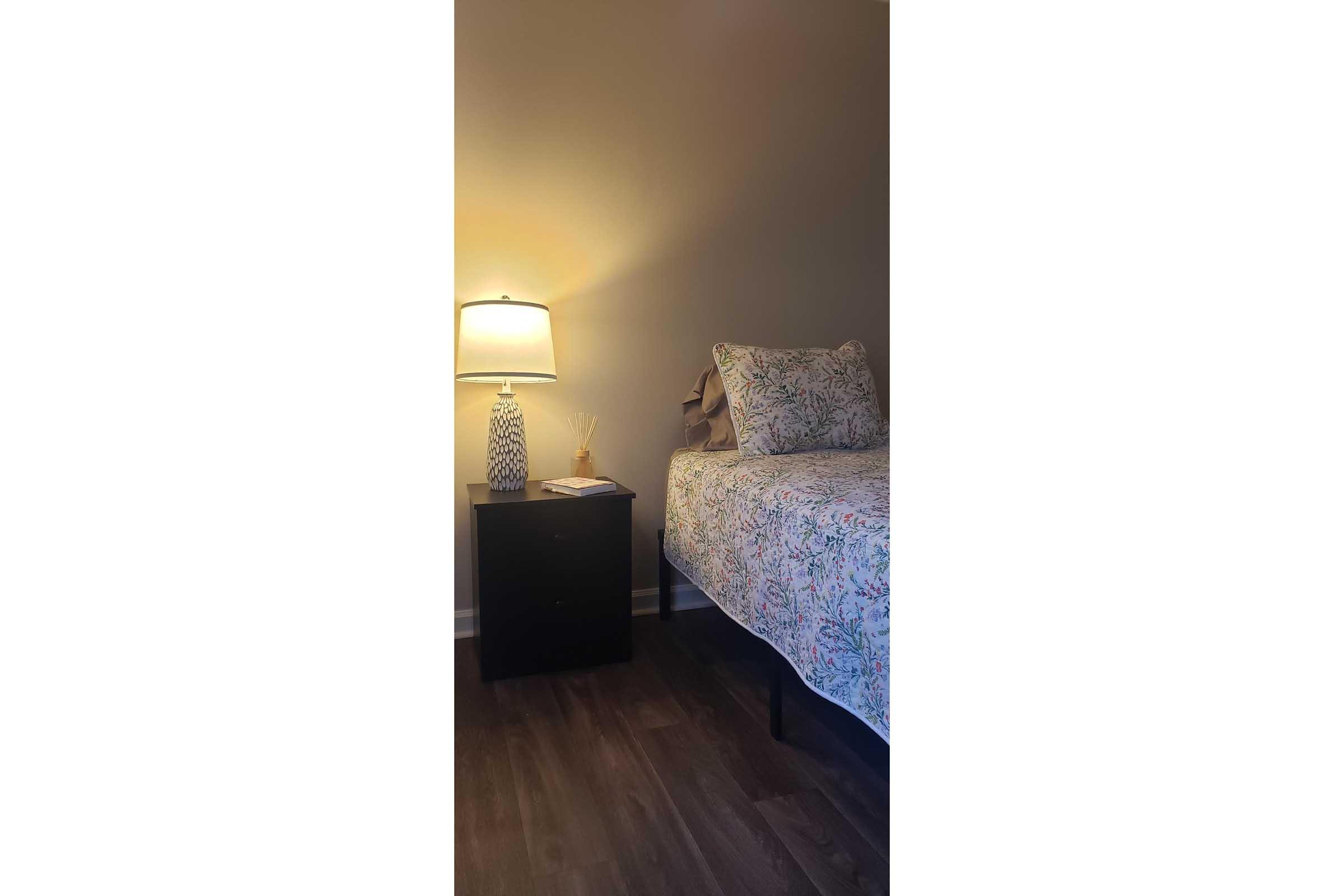 A cozy bedroom corner featuring a bed with a floral-patterned quilt, a bedside table with a lamp, and a book on top. Soft lighting creates a warm atmosphere, with wooden flooring visible in the foreground.