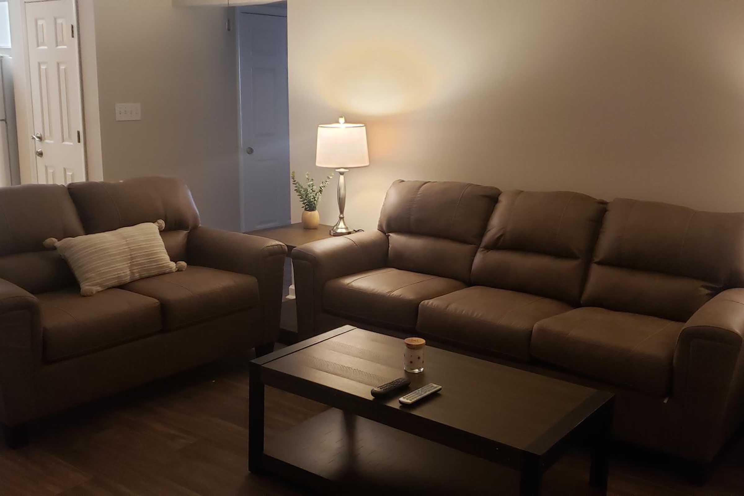 A cozy living room featuring two brown leather sofas, a coffee table with a remote control and a candle, and a lamp providing warm light. There is a small plant on the table, and a door in the background indicates access to another room. The floor is wood, adding to the inviting atmosphere.