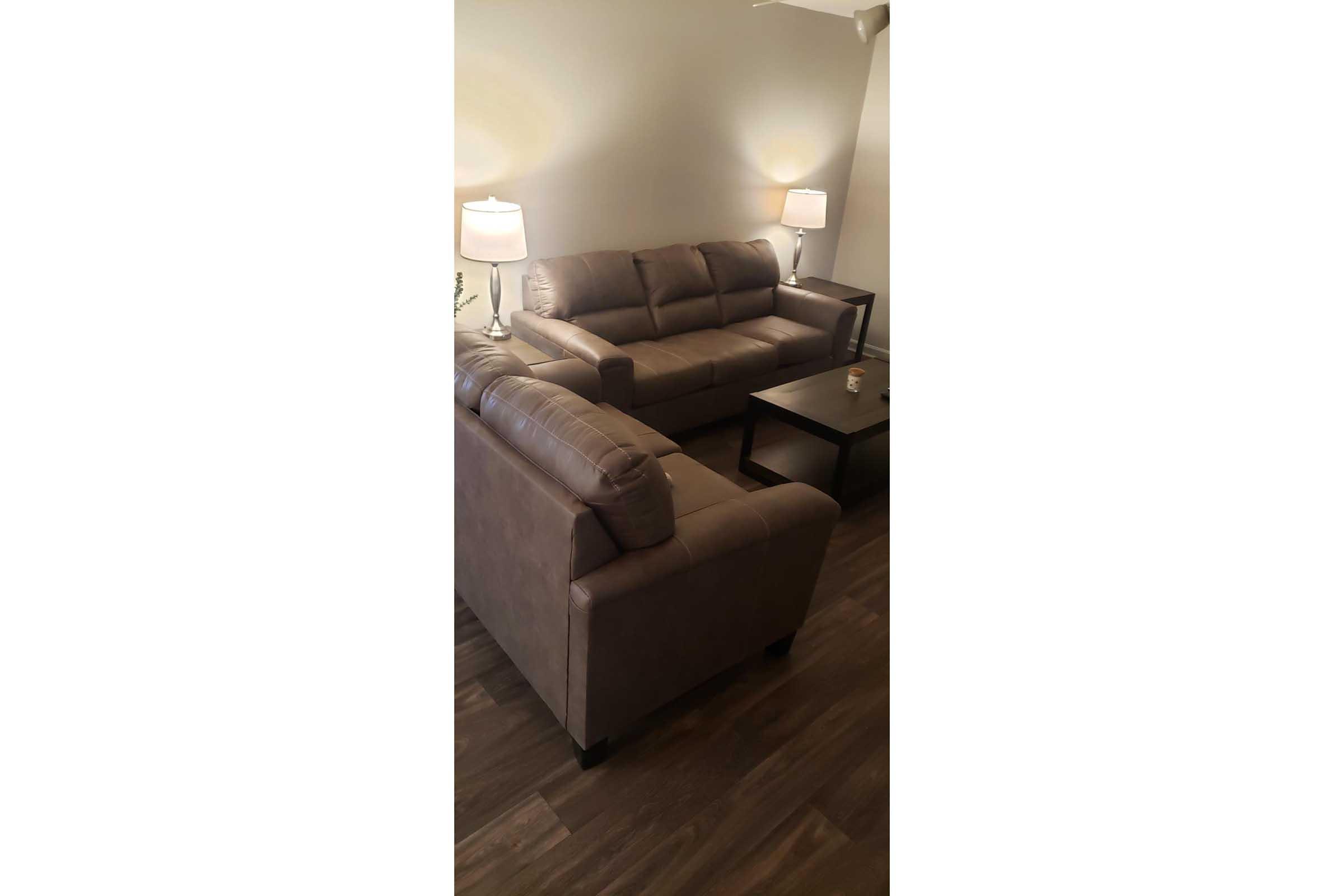 A cozy living room interior featuring a brown leather couch and loveseat arranged around a small coffee table. Two lamps on either side create a warm ambiance, and the flooring is a rich, dark wood. The walls are light-colored, enhancing the room's inviting atmosphere.