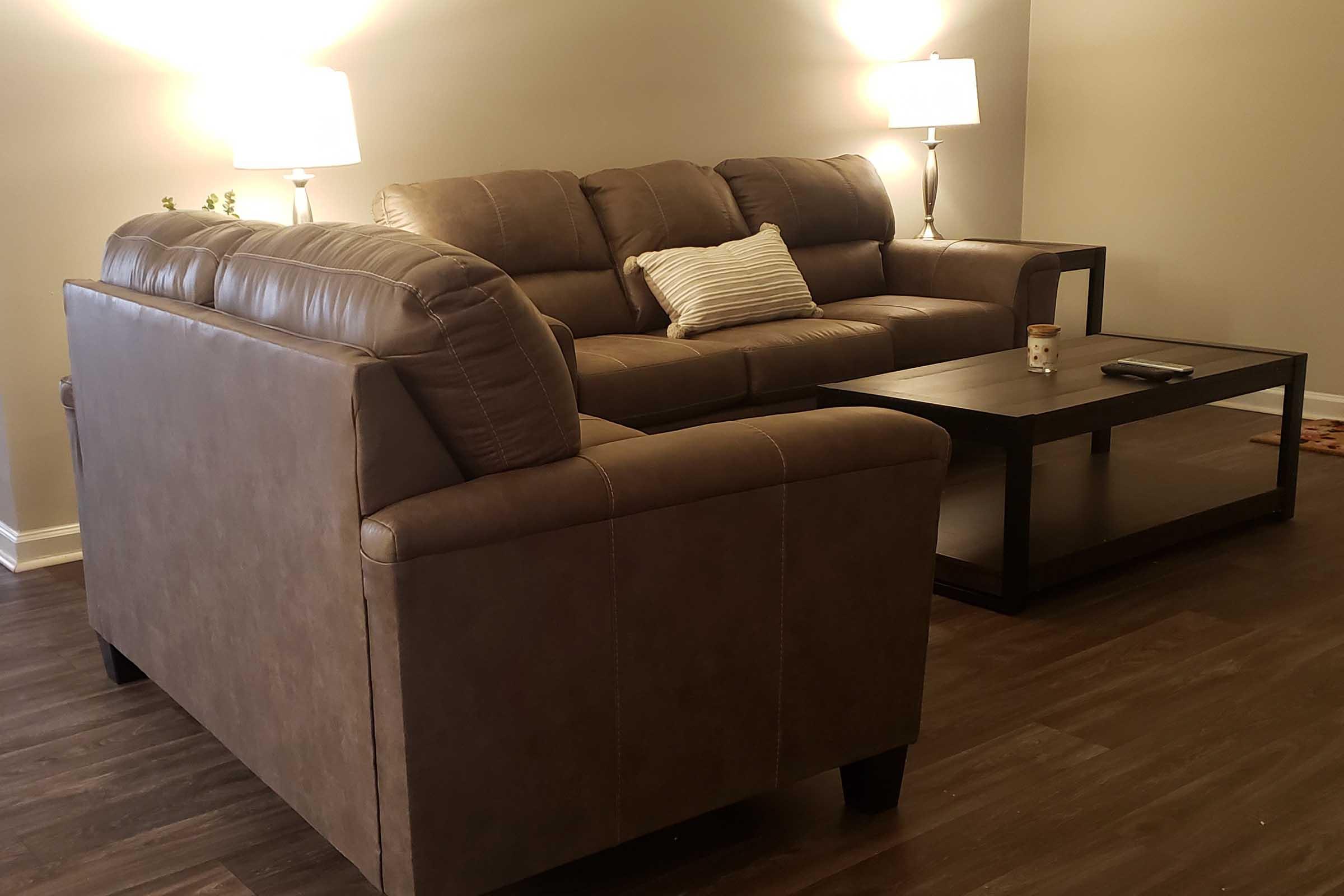 A cozy living room featuring a brown leather sofa and an armchair, both positioned around a wooden coffee table. Two lamps illuminate the space, creating a warm ambiance. A small plant is visible in the background, adding a touch of greenery to the decor. The flooring is a dark wood finish.