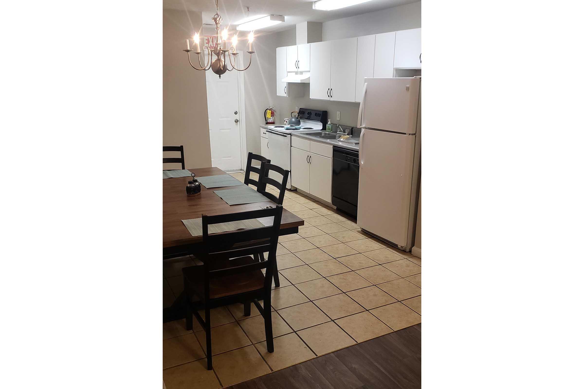 A spacious kitchen with white cabinets and appliances. The room features a wooden dining table with black chairs, a chandelier overhead, and a tiled floor. A door leads to the outside, and various kitchen items are visible on the countertop and stove.