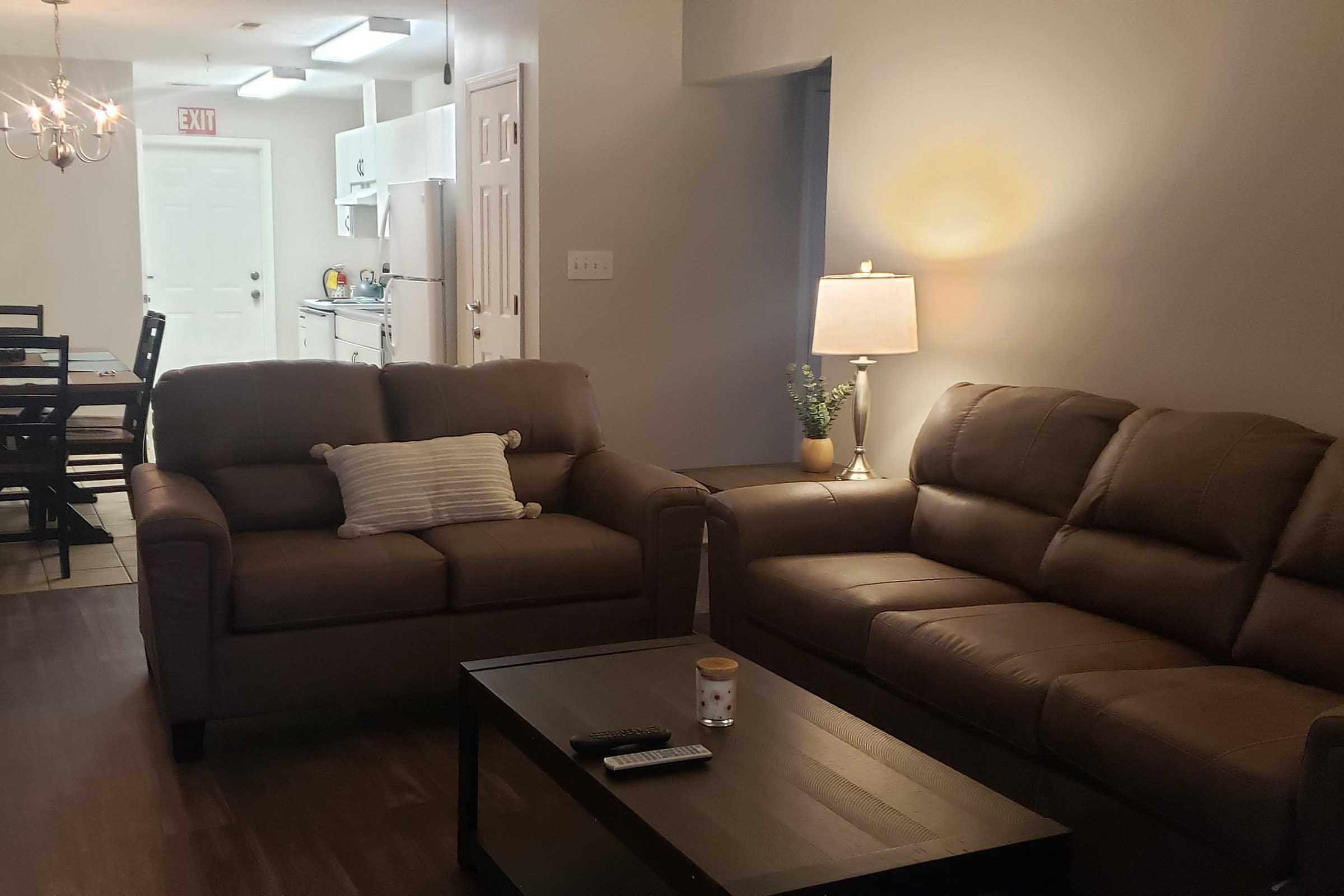 A cozy living room featuring two brown leather sofas, a coffee table with a remote and a candle on it. A lamp illuminates the space, and in the background, a dining area and kitchen can be seen. The walls are light-colored, and there is a small plant beside the lamp.