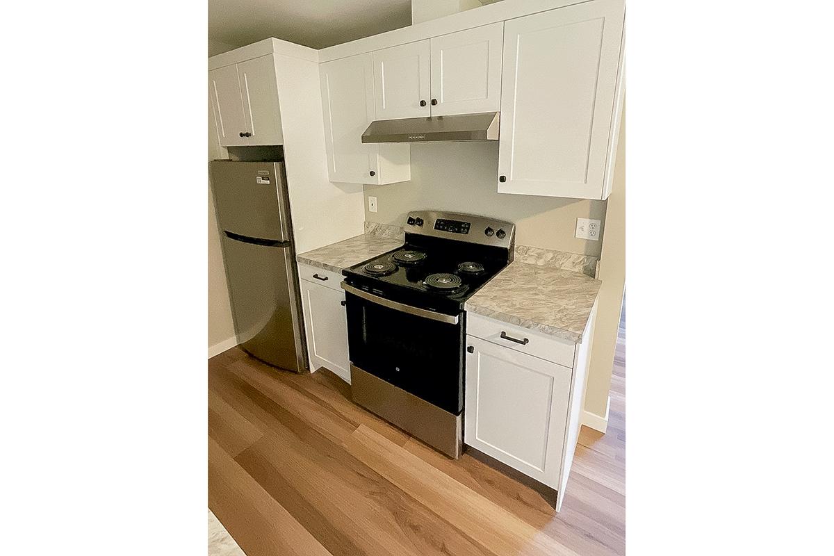 Modern kitchen featuring white cabinetry, stainless steel refrigerator, and a black gas stove with a hood. The countertops have a marbled design, and the flooring is wood. The kitchen appears clean and well-organized, with ample space and natural lighting.
